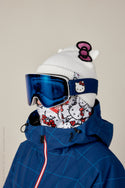 A person in a blue ski jacket sports a white Hello Kitty beanie, Hello Kitty neck gaiter, and blue 686 SMARTY® Magnetic Goggle, completing their look with several Hello Kitty accessories and designs.