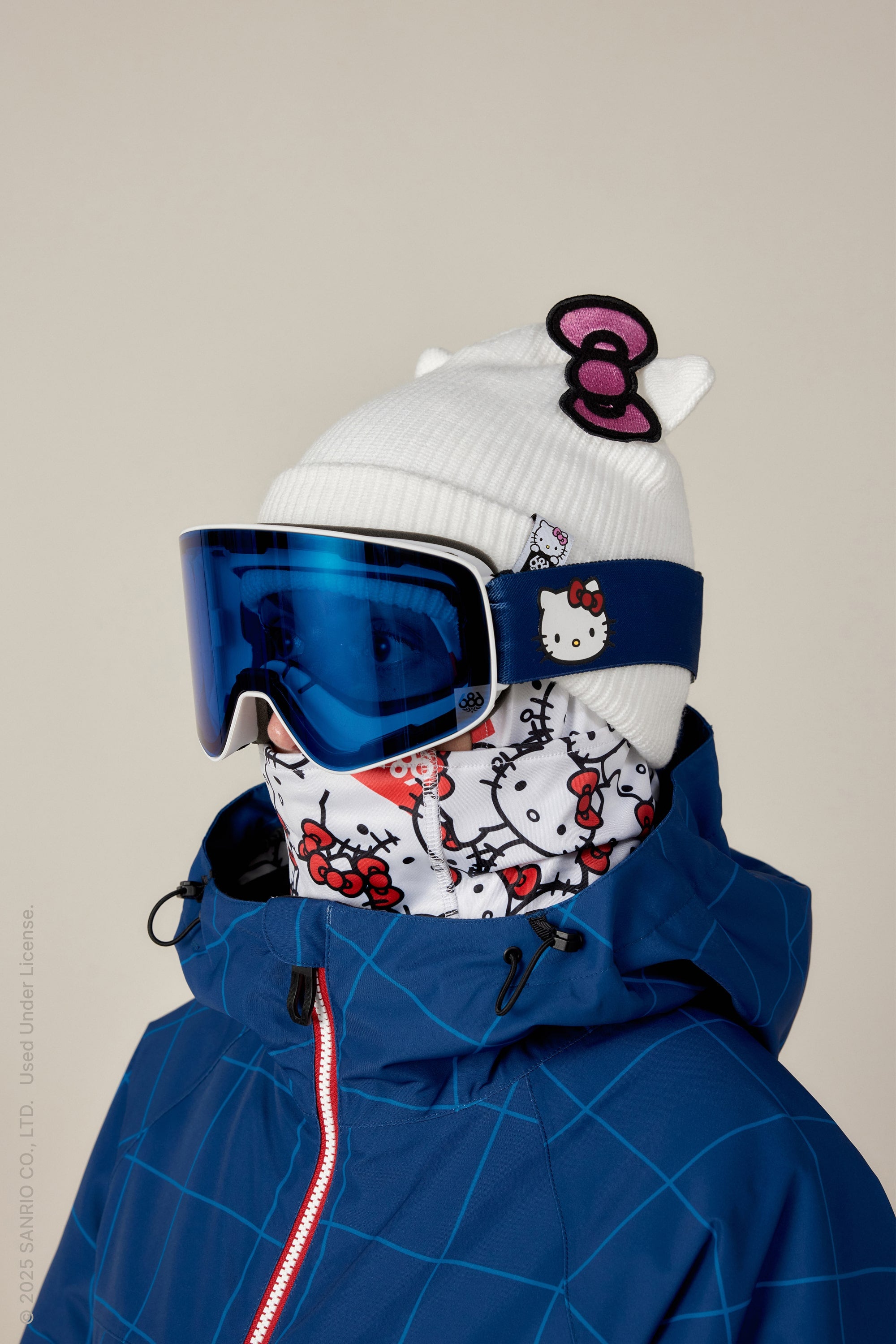 A person in a blue ski jacket sports a white Hello Kitty beanie, Hello Kitty neck gaiter, and blue 686 SMARTY® Magnetic Goggle, completing their look with several Hello Kitty accessories and designs.