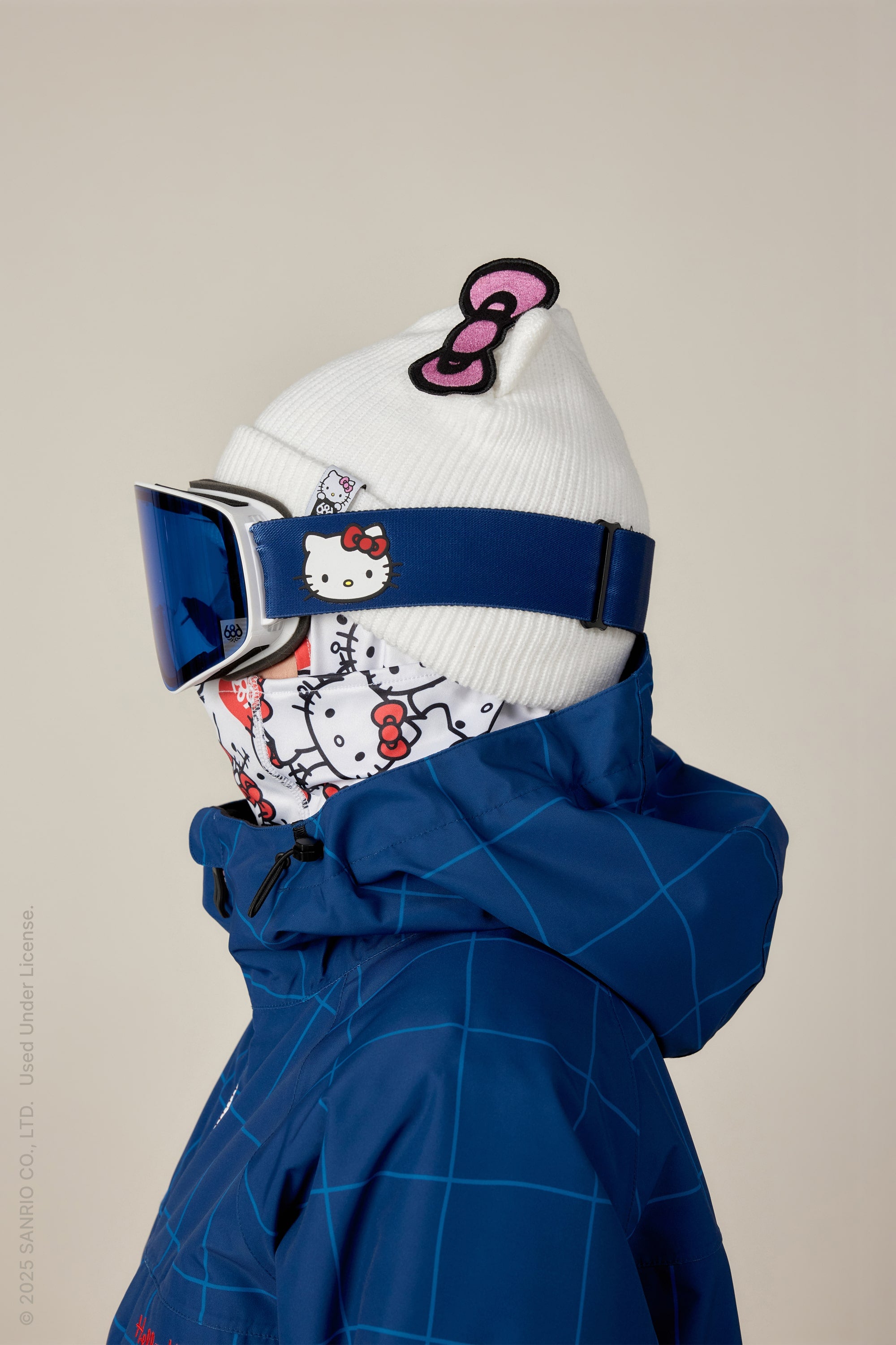 A person in a 686 SMARTY Magnetic Goggle and a blue jacket wears a white beanie with a pink Hello Kitty bow and a matching Hello Kitty face mask, both featuring the character’s design, against a plain background.