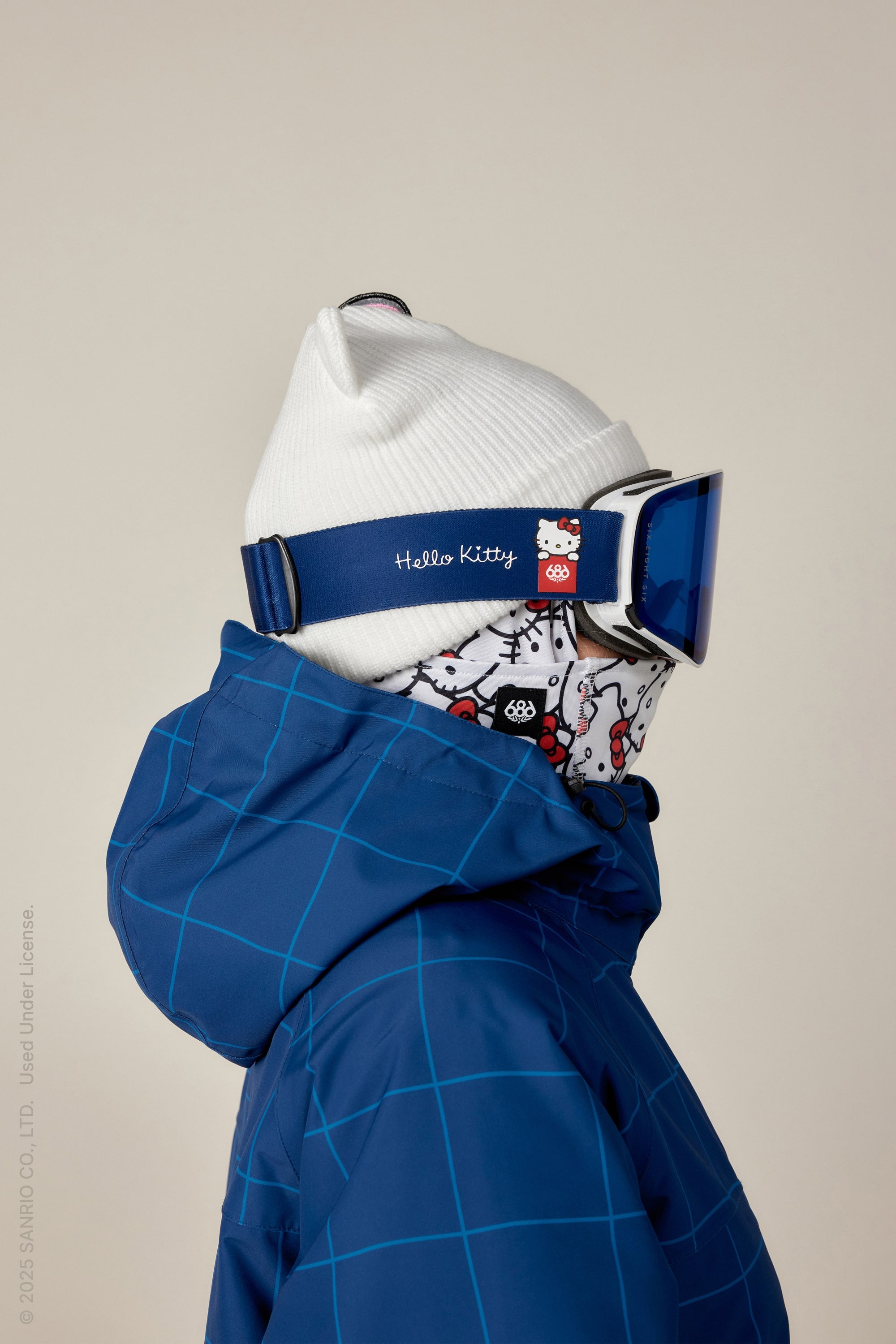 Person in a white beanie, blue ski jacket, and 686 SMARTY Magnetic Goggle by 686 with a Hello Kitty patterned face covering, shown in profile against a neutral background.