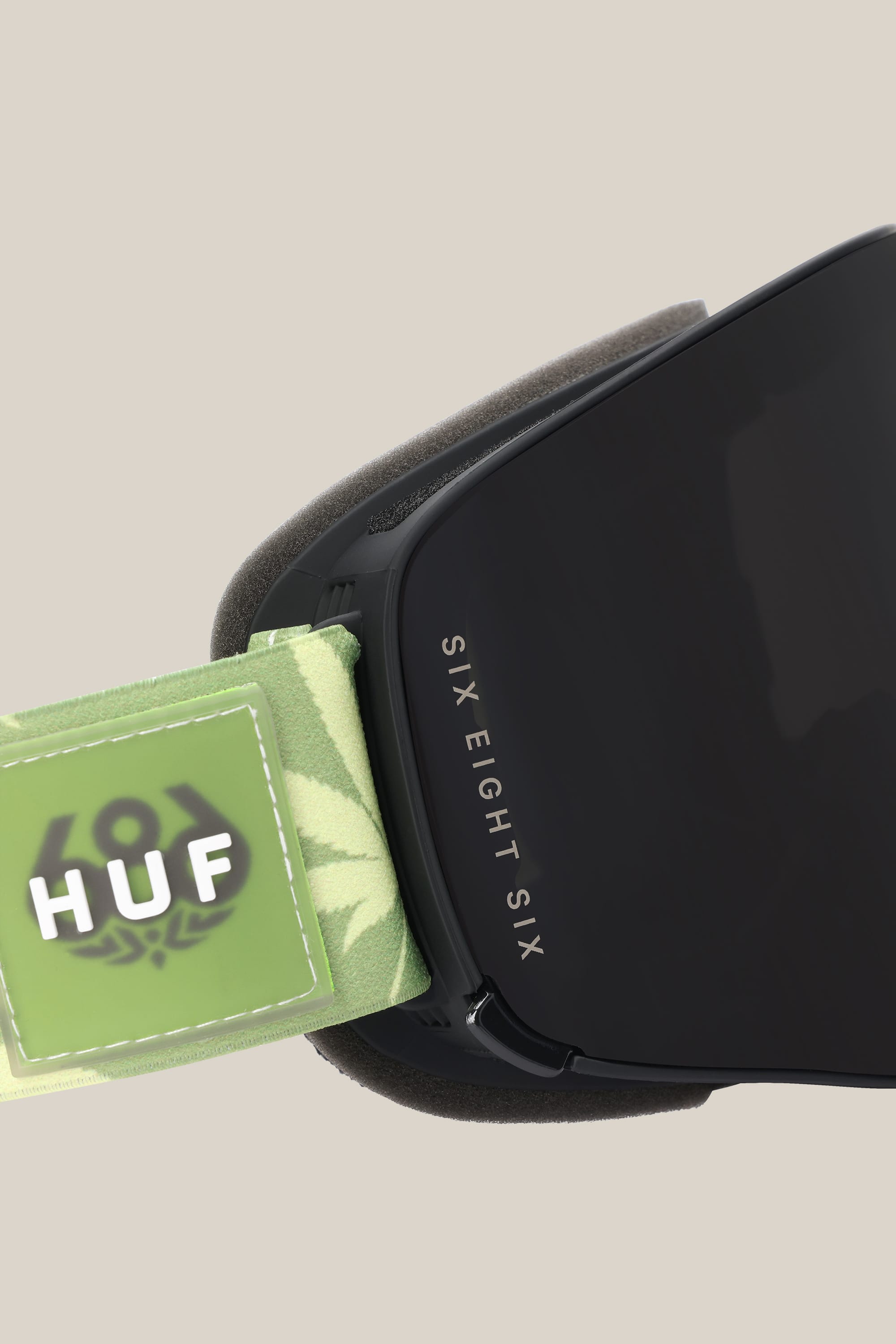 Close-up of the 686 SMARTY® Magnetic Goggle in black, featuring SIX EIGHT SIX on the frame and a green leafy-patterned strap with the HUF logo, set against a plain beige background.