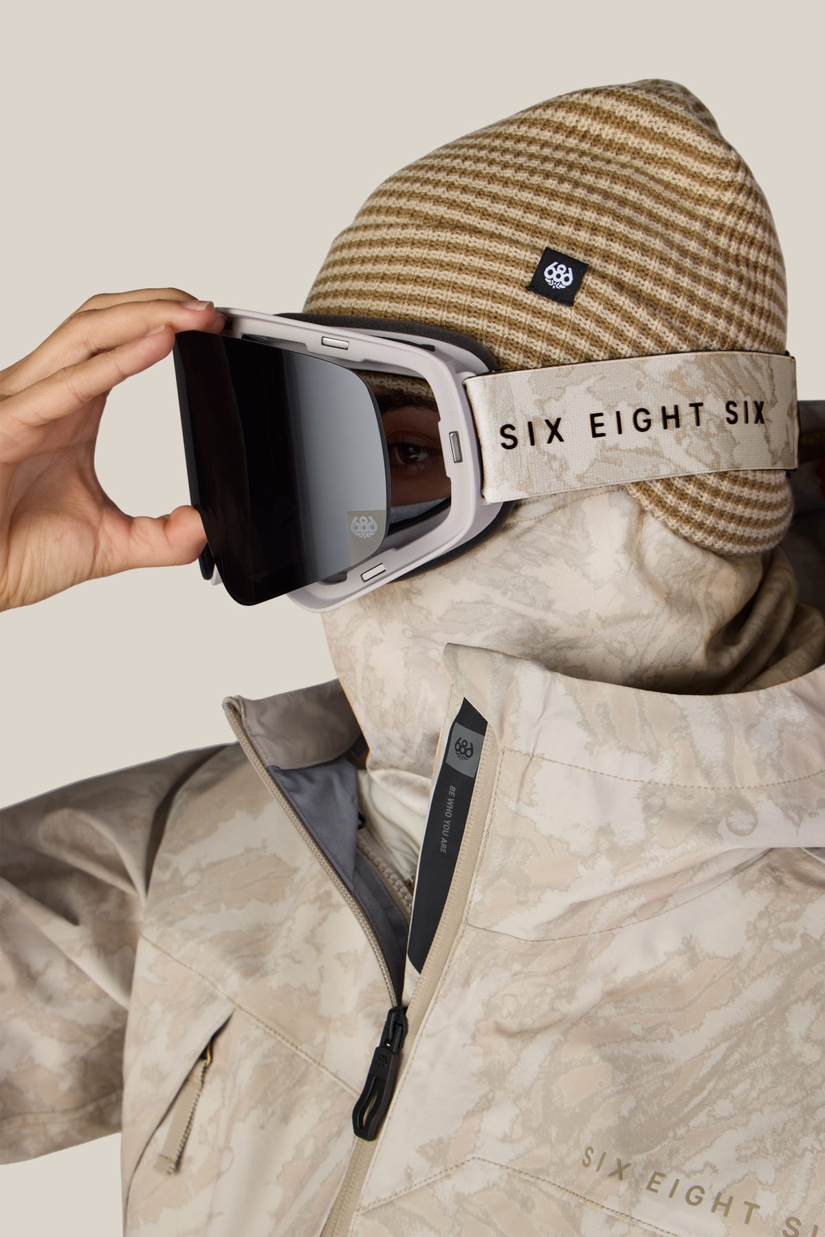 A person wearing a beige patterned winter jacket, matching hat, face covering, and large ski goggles labeled 686 holds the 686 SMARTY® Magnetic Goggle with one hand against a plain background.