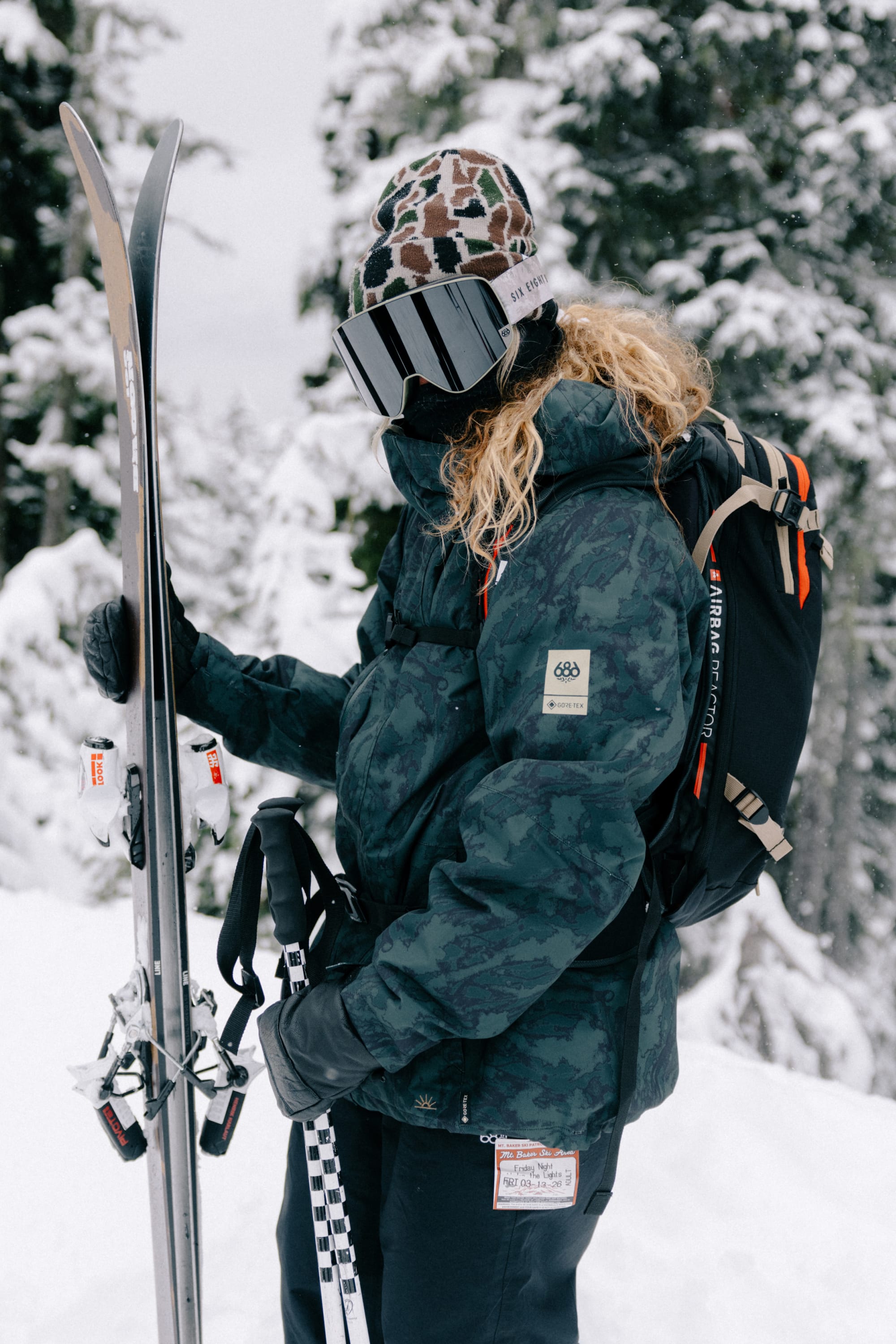 Wearing the 686 SMARTY® Magnetic Goggle by 686, a person in winter gear with a backpack holds a pair of skis and ski poles while standing in a snowy forest.
