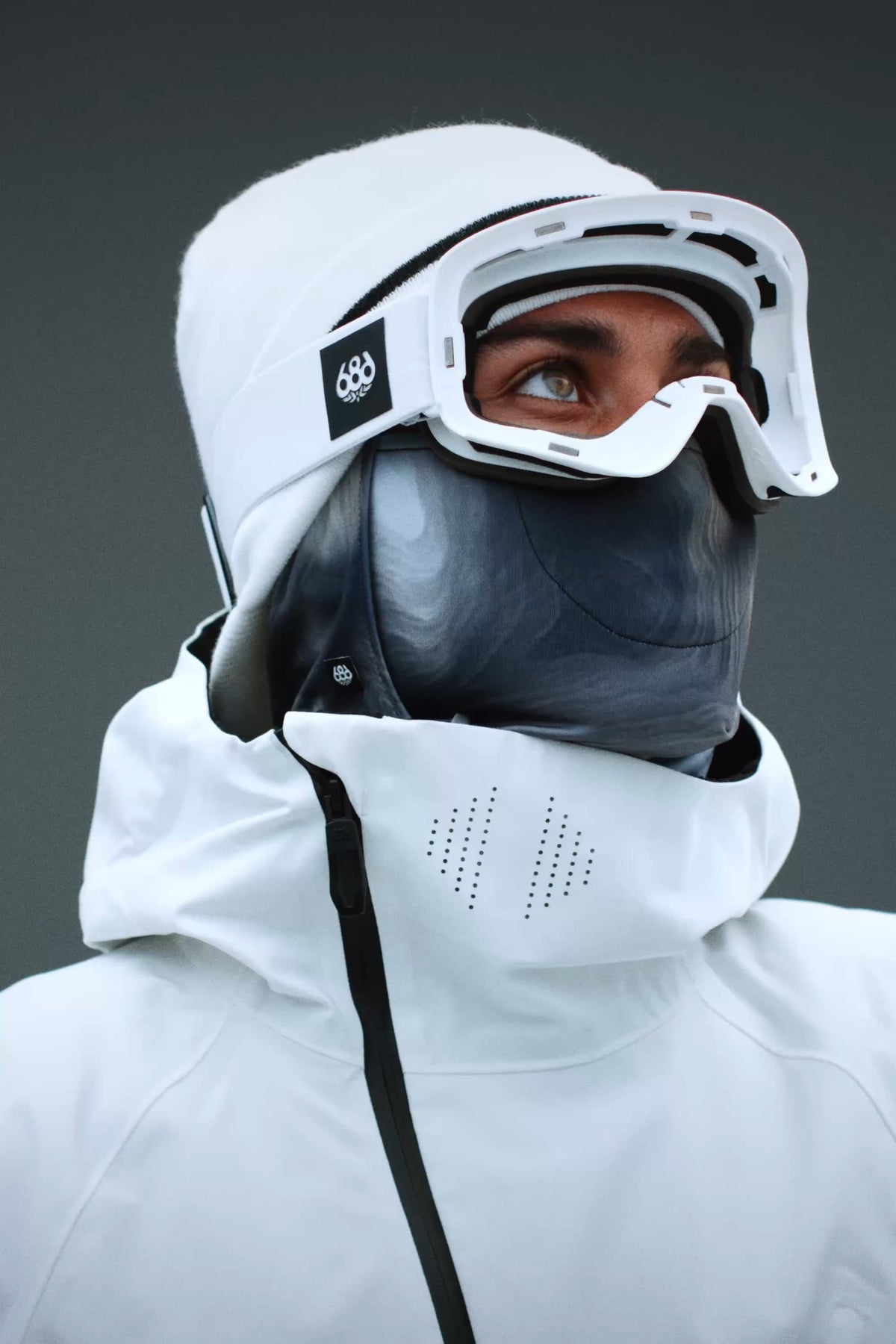 A person wearing a white jacket with a hood and the 686 SMARTY® Magnetic Goggle by 686 looks upward. Their face is mostly covered by a dark mask, and the background is plain gray.