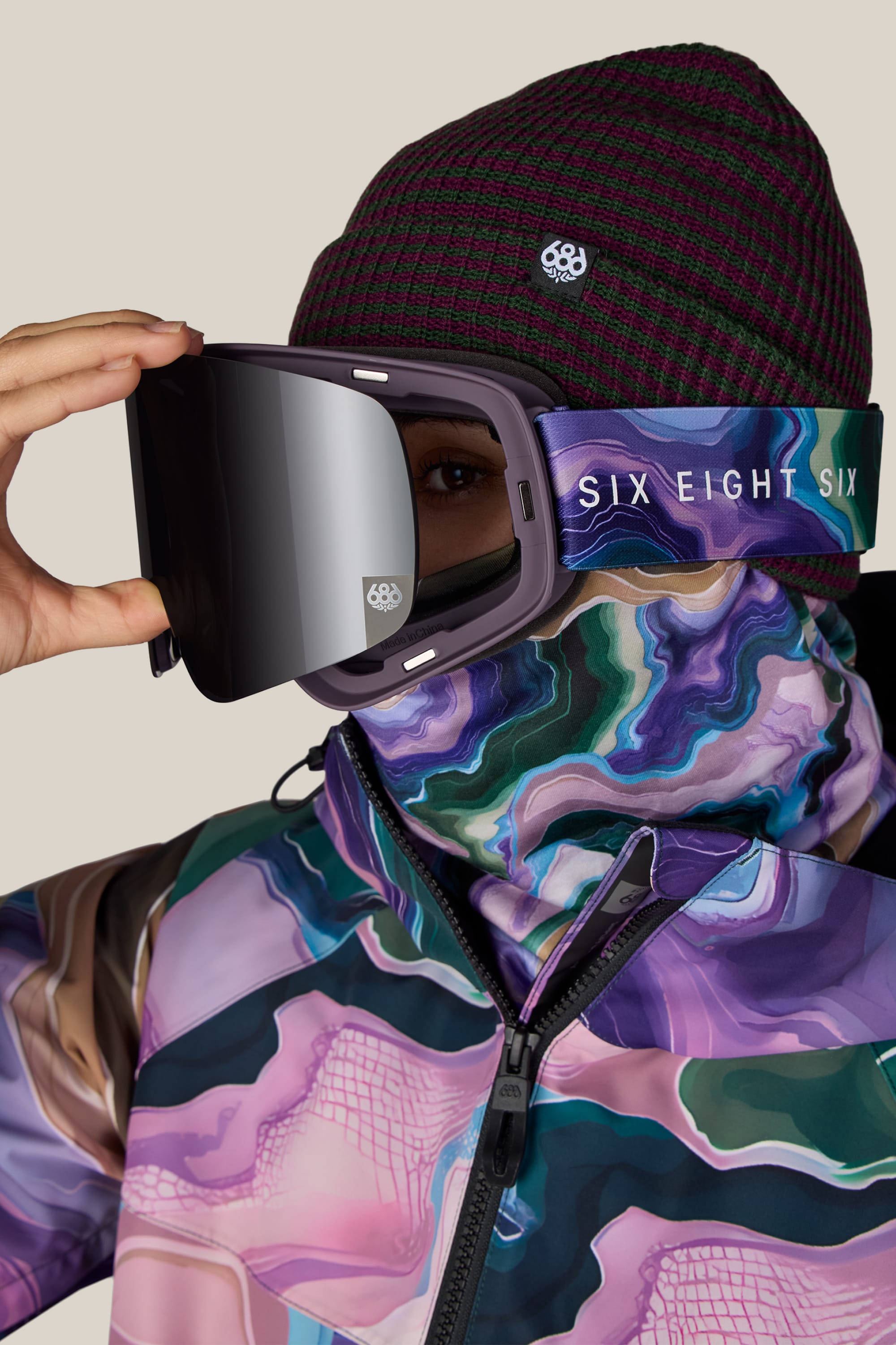A person wearing the 686 SMARTY® Magnetic Goggle with a branded 686 strap, along with a maroon patterned beanie and a colorful, abstract-patterned jacket and neck gaiter, set against a plain beige background.