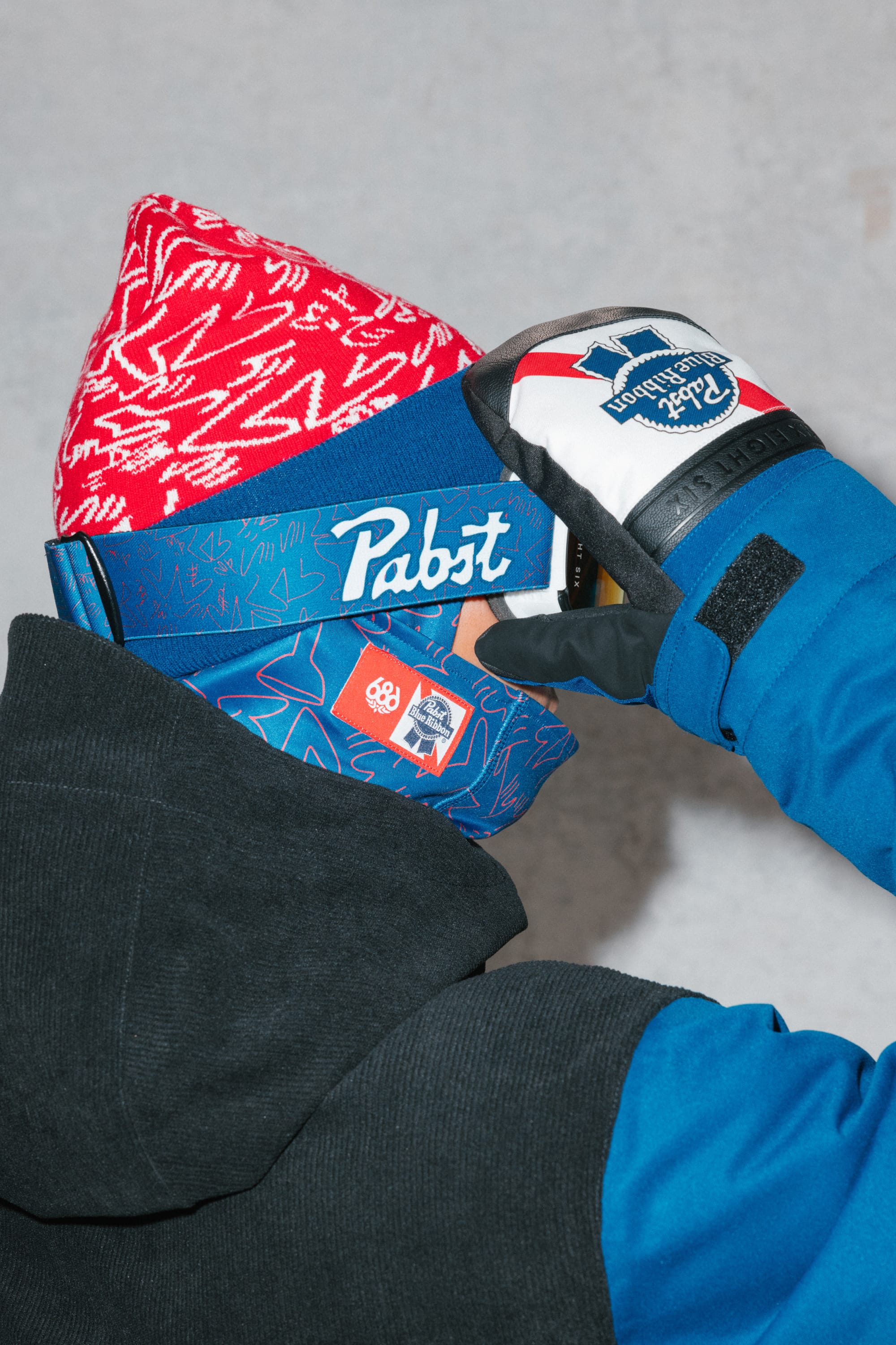 A person in a red knit hat, blue jacket, and patterned neck gaiter holds up a gloved hand with a Pabst Blue Ribbon logo near their face, wearing 686 SMARTY® Magnetic Goggles by 686, standing against a plain light gray background.