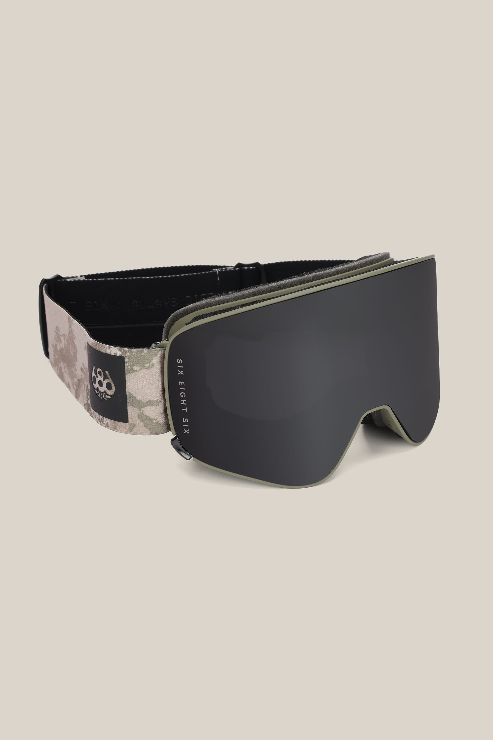 The 686 SMARTY® Magnetic Goggle features a minimalist design with a wide dark reflective lens, beige and gray camo adjustable strap, small white logo patch, and is compatible with interchangeable lenses.