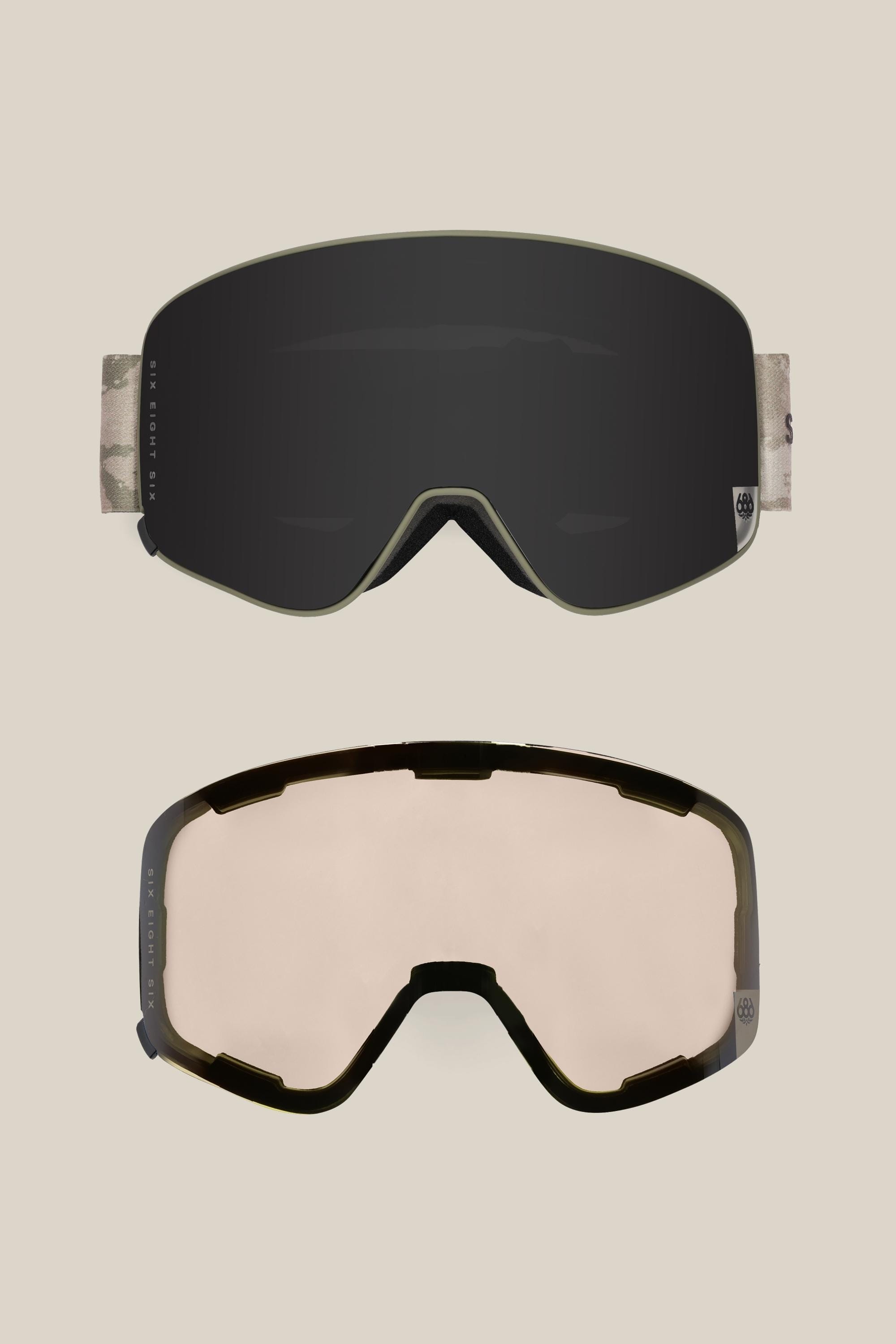 Two 686 SMARTY® Magnetic Goggles by 686 are shown on a beige background: one has a dark mirrored lens with camo-patterned strap, the other features a clear lens and dark frame; both offer easy interchangeable lenses.