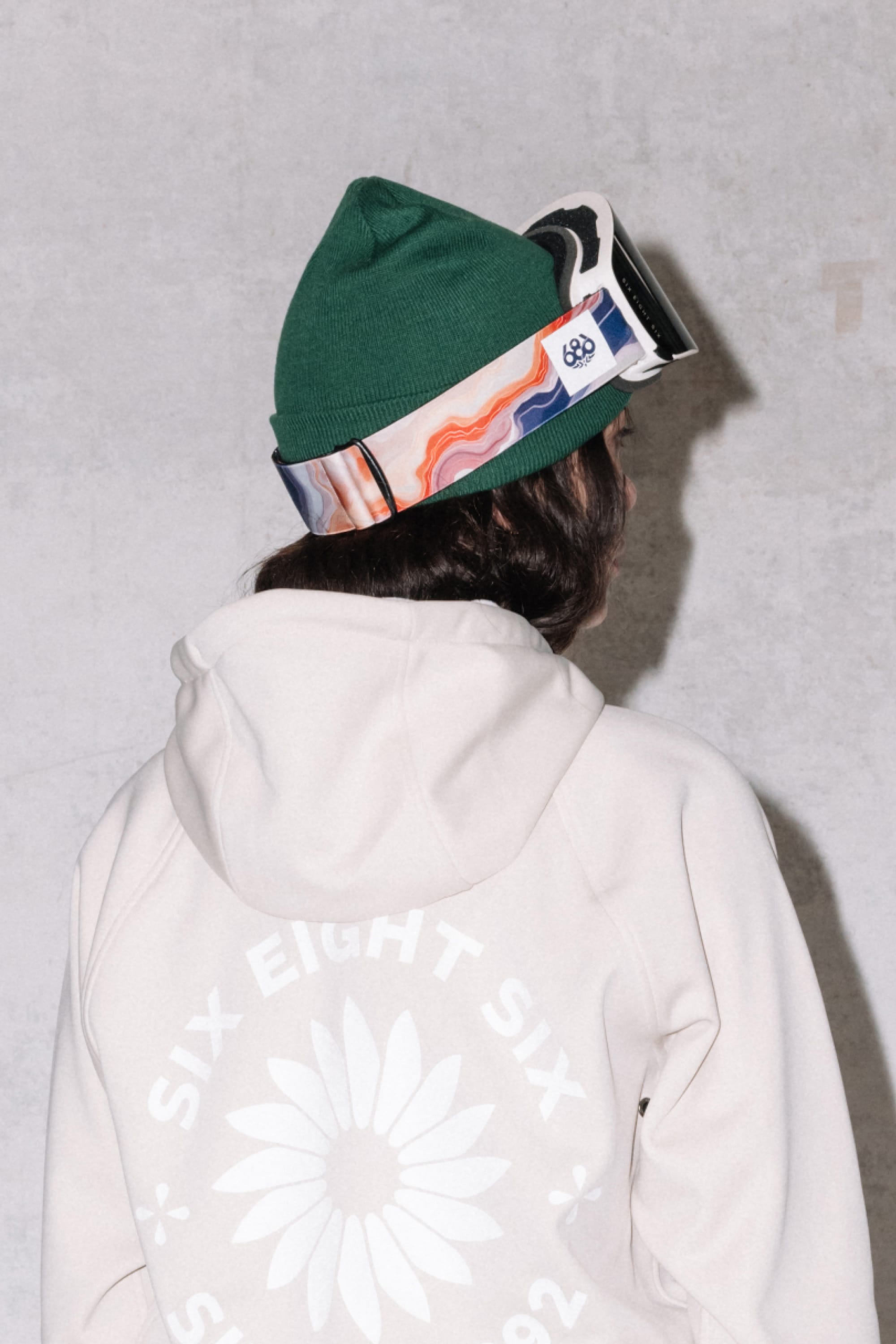 A person in a green beanie wears 686 SMARTY® Magnetic Goggles, seen from behind in a light hoodie featuring a large daisy and "SIX EIGHT SIX SINCE 1992" printed on the back.