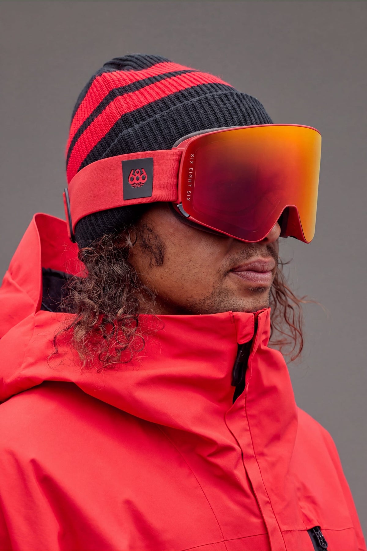 A person with long, curly hair partially visible under a red and black beanie wears the 686 SMARTY® Magnetic Goggle by 686 and a red jacket, facing slightly to the right against a plain gray background.