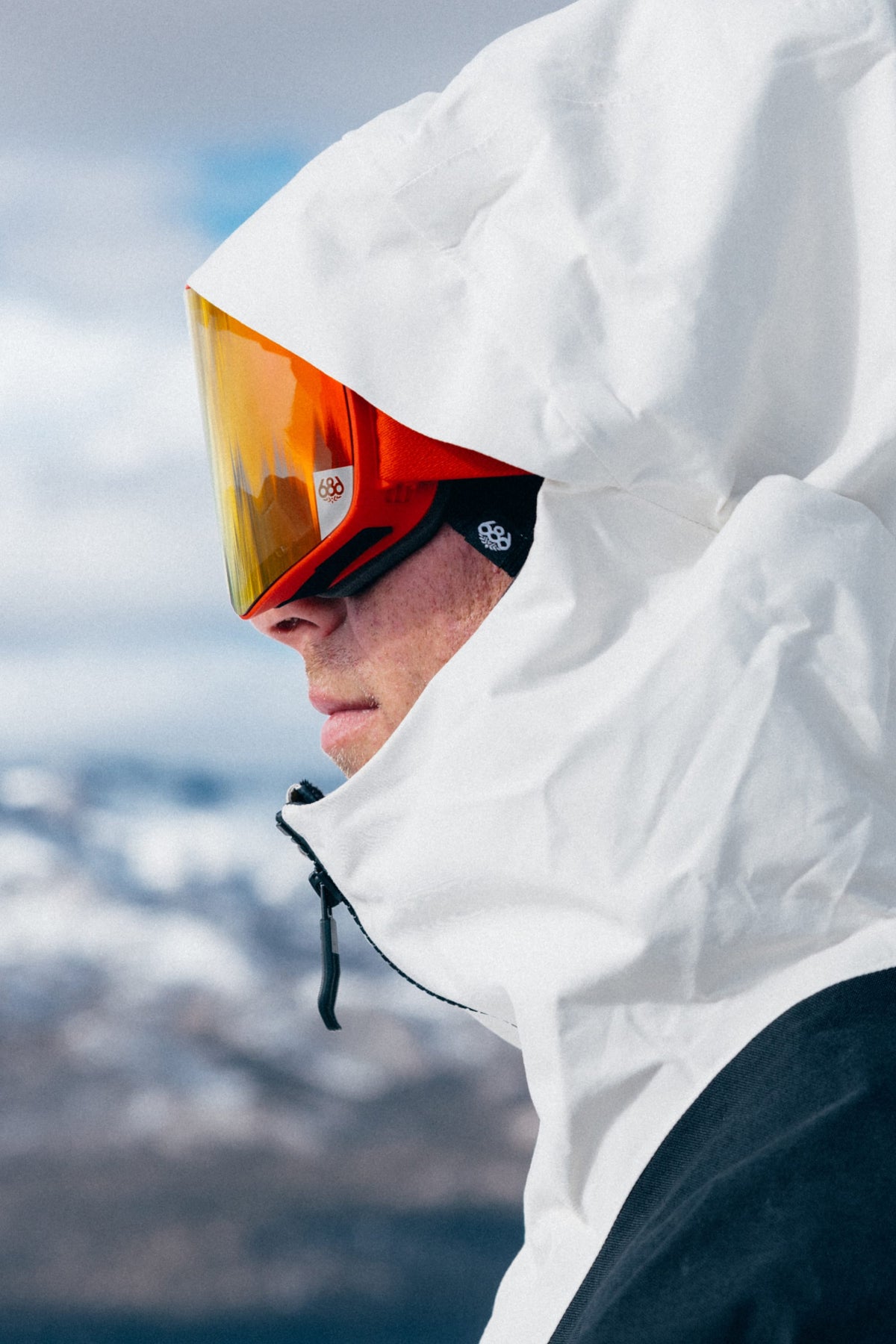 A person wearing a white ski jacket, red helmet, and 686 SMARTY® Magnetic Goggle by 686 stands outdoors with snowy mountains blurred in the background.