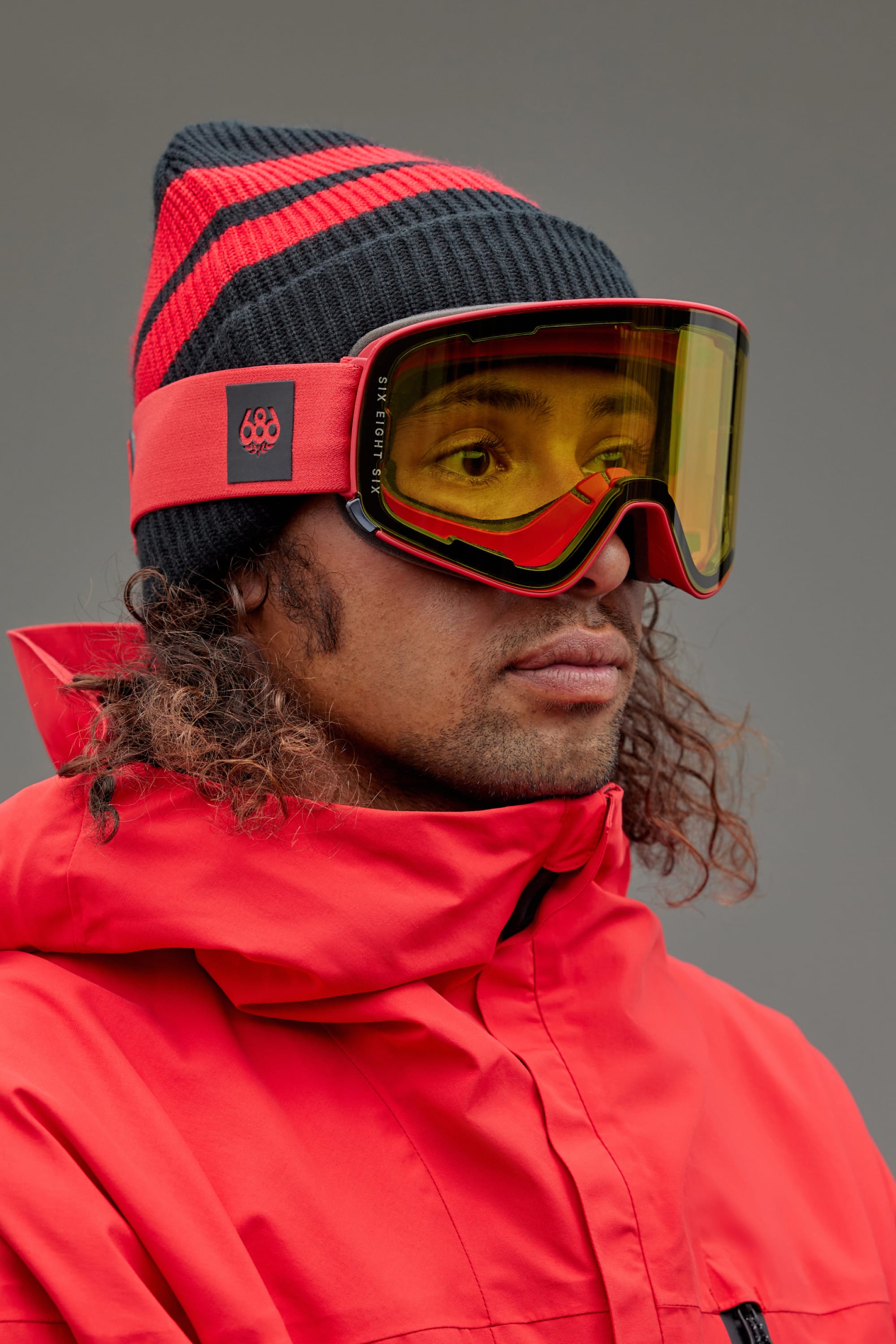 A person in a red and black striped beanie, yellow-tinted 686 SMARTY® Magnetic Goggle with red frame, and a red winter jacket looks to the side against a plain gray background.