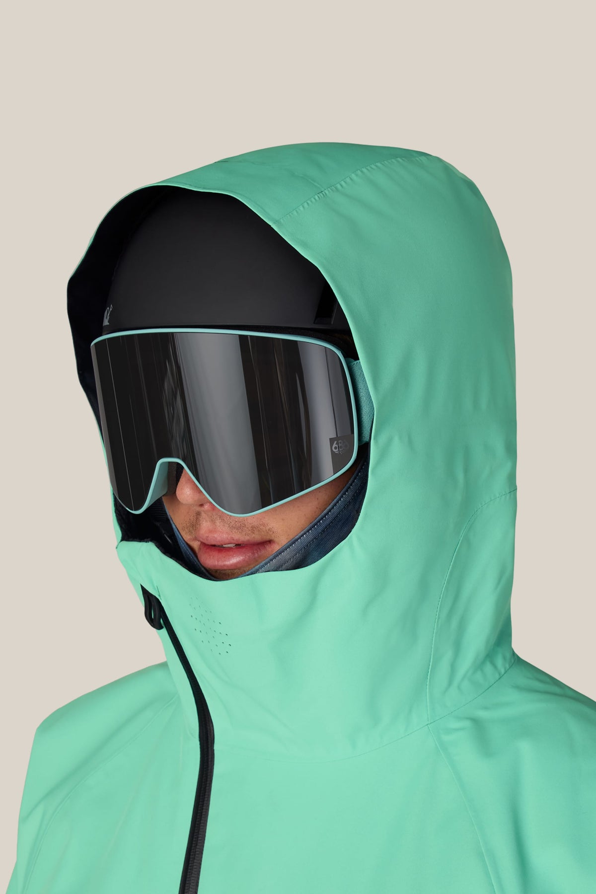 A person wearing a mint green hooded jacket, a black ski helmet, and large 686 SMARTY® Magnetic Goggles by 686, facing left against a neutral beige background.