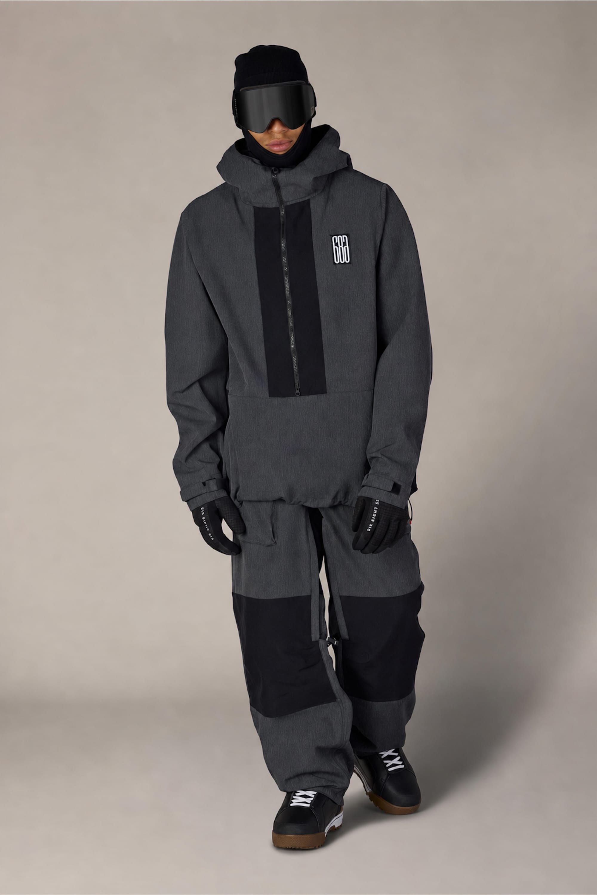 A person dressed in the 686 Men’s Ghost™ 2.5L Anorak by 686, paired with baggy pants, gloves, black boots, a beanie, and ski goggles, stands against a plain light background.
