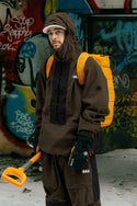 A person in the 686 Men's Ghost™ 2.5L Anorak and matching pants holds an orange snow shovel, wearing an orange backpack, in front of a graffiti-covered wall with snow blanketing the ground.