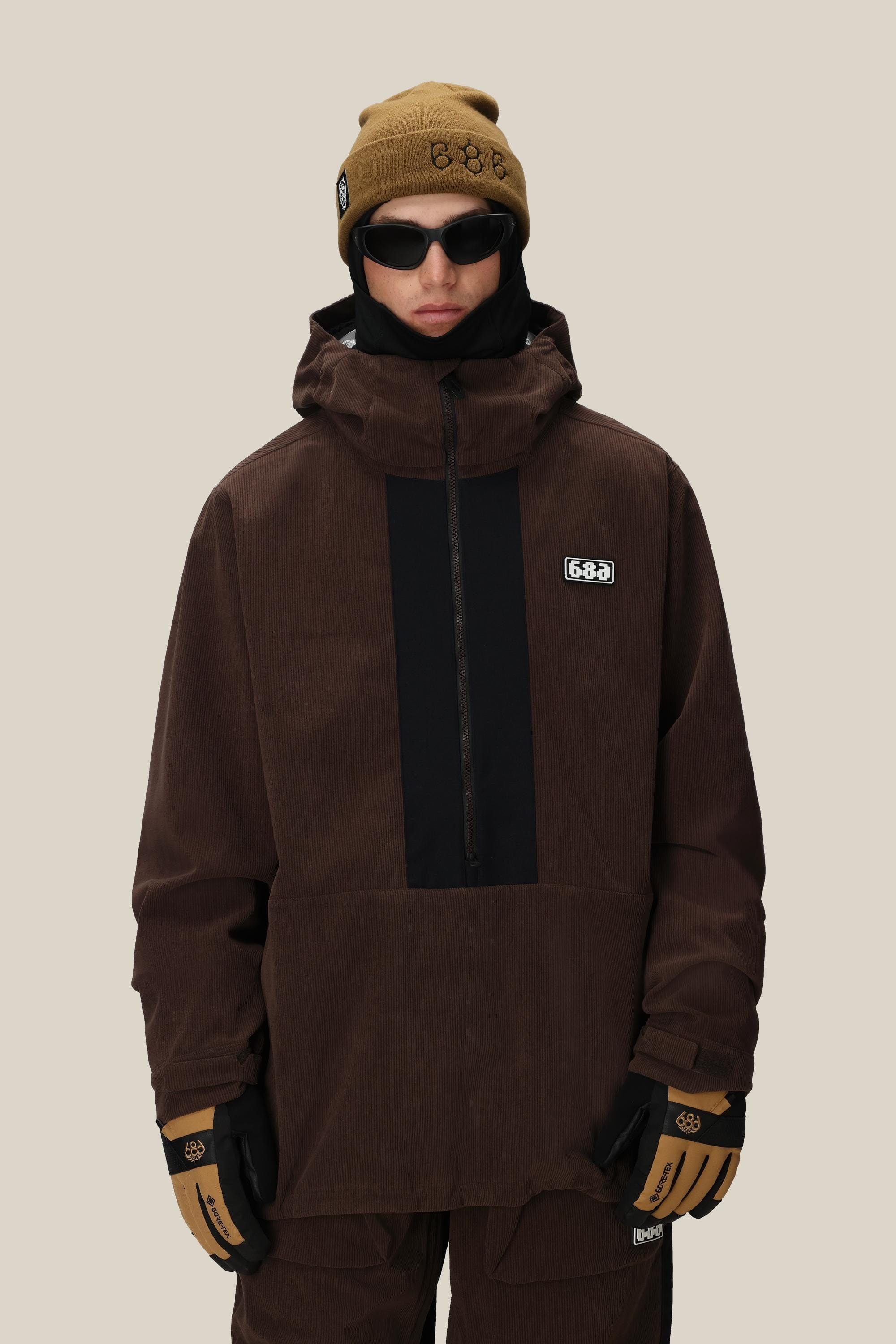 A person wearing the 686 Men's Ghost™ 2.5L Anorak—a rugged brown nylon hooded jacket with black accents—plus matching pants, tan gloves, a tan beanie, and black sunglasses stands against a plain beige background in a modern shorter fit.