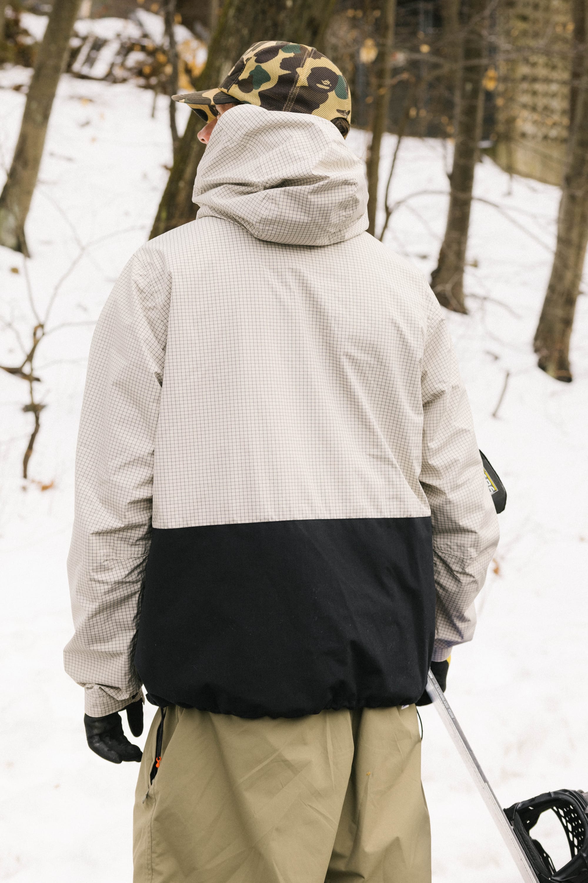 Wearing the 686 Men's Ghost™ 2.5L Anorak, a person in a camouflage hat stands in a snowy forest, facing away from the camera and holding ski poles at their side.