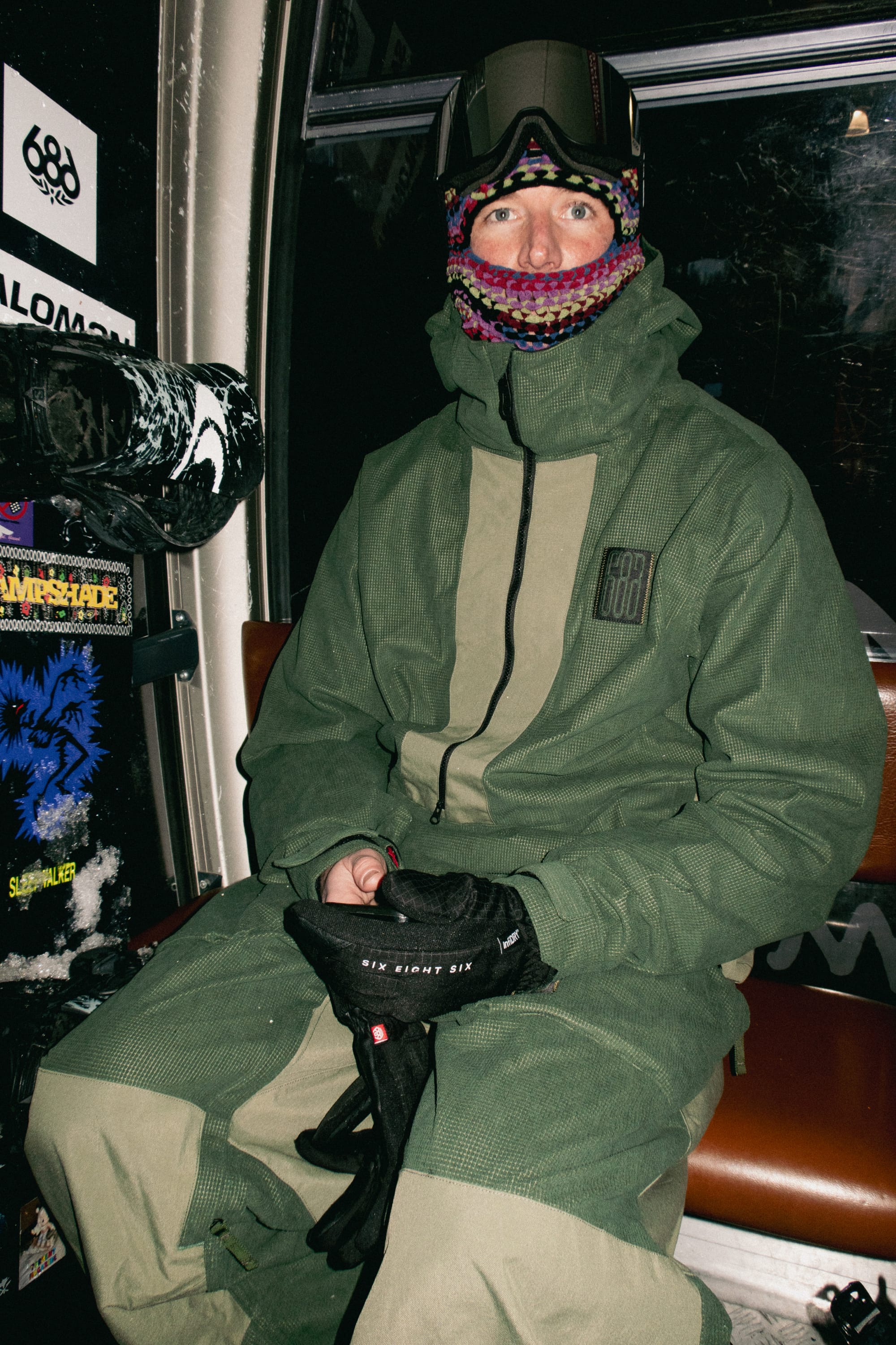 Wearing the 686 Men’s Ghost™ 2.5L Anorak, a person sits on a bench inside a sticker-covered ski gondola, sporting colorful face mask, ski goggles, and holding black gloves.