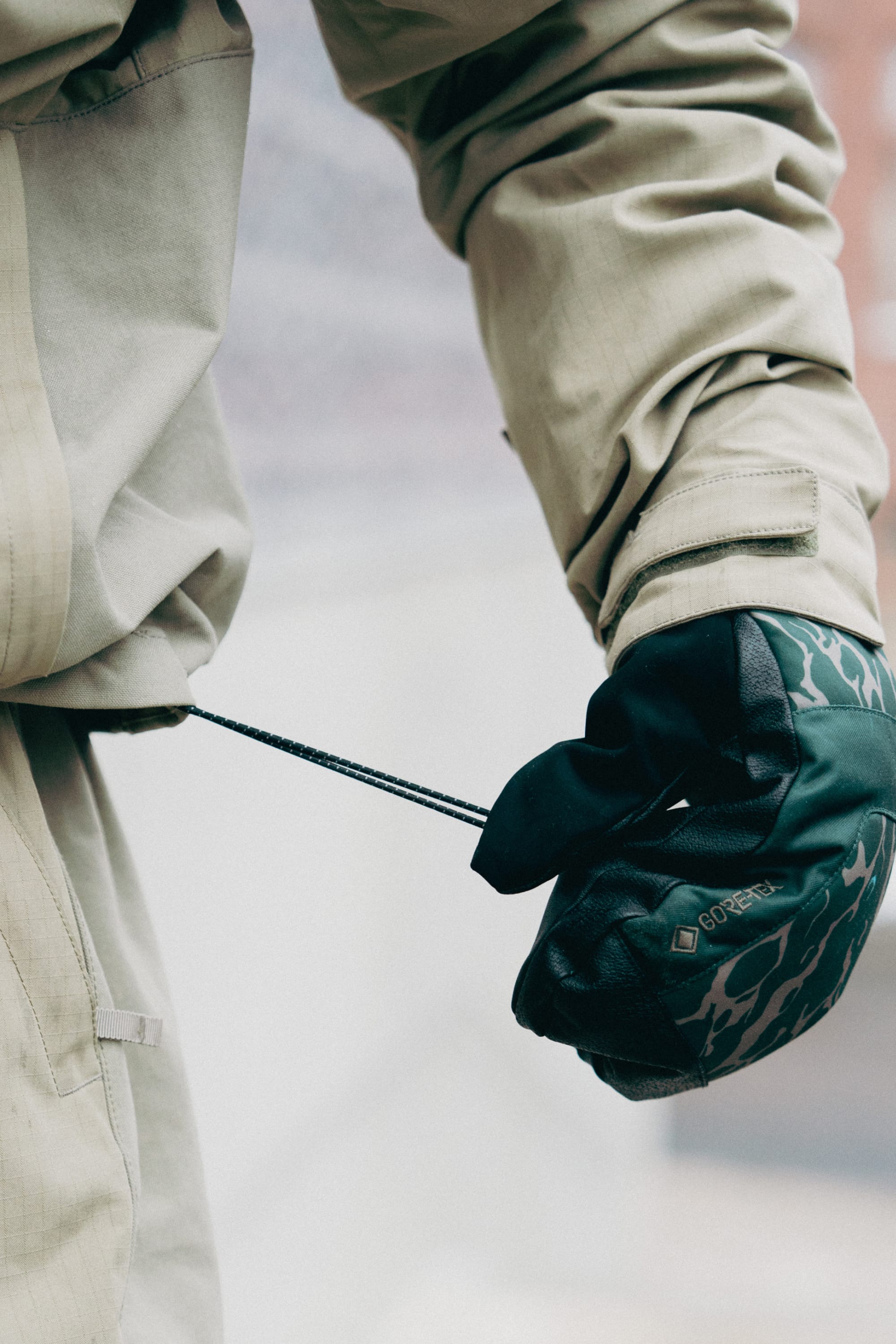 A person in the 686 Men's Ghostª 2.5L Anorak tightens the adjustable waist cord. They're also wearing dark patterned gloves, and the background is blurred.