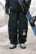 A person in 686 Ghost™ 2.5L Pants and boots stands on a snowy path, holding a snowboard with black bindings upright. Patches and graphics accent the pants against a wintry, wooded backdrop.