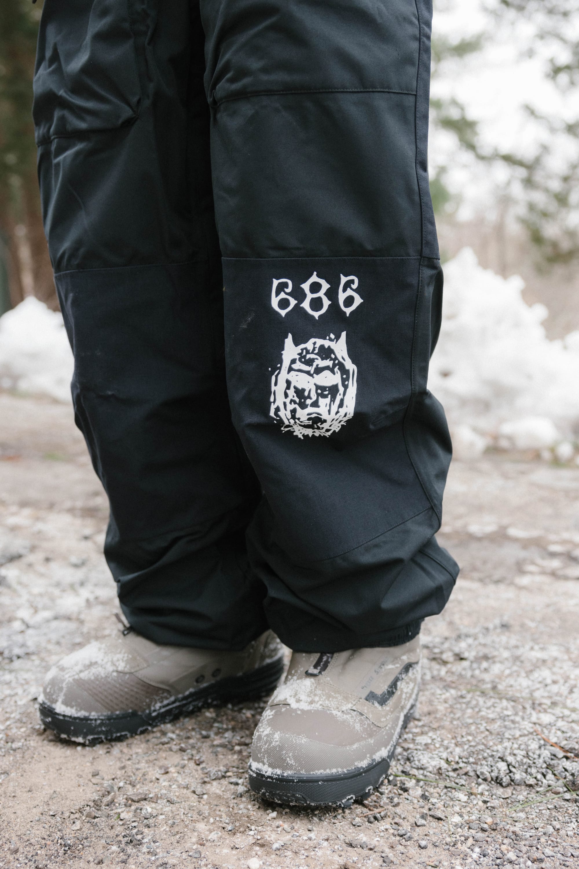 A person wearing black 686 Ghost™ 2.5L Pants stands on a snowy path in gray snow boots, with snow-dusted trees and a wintry landscape in the background.