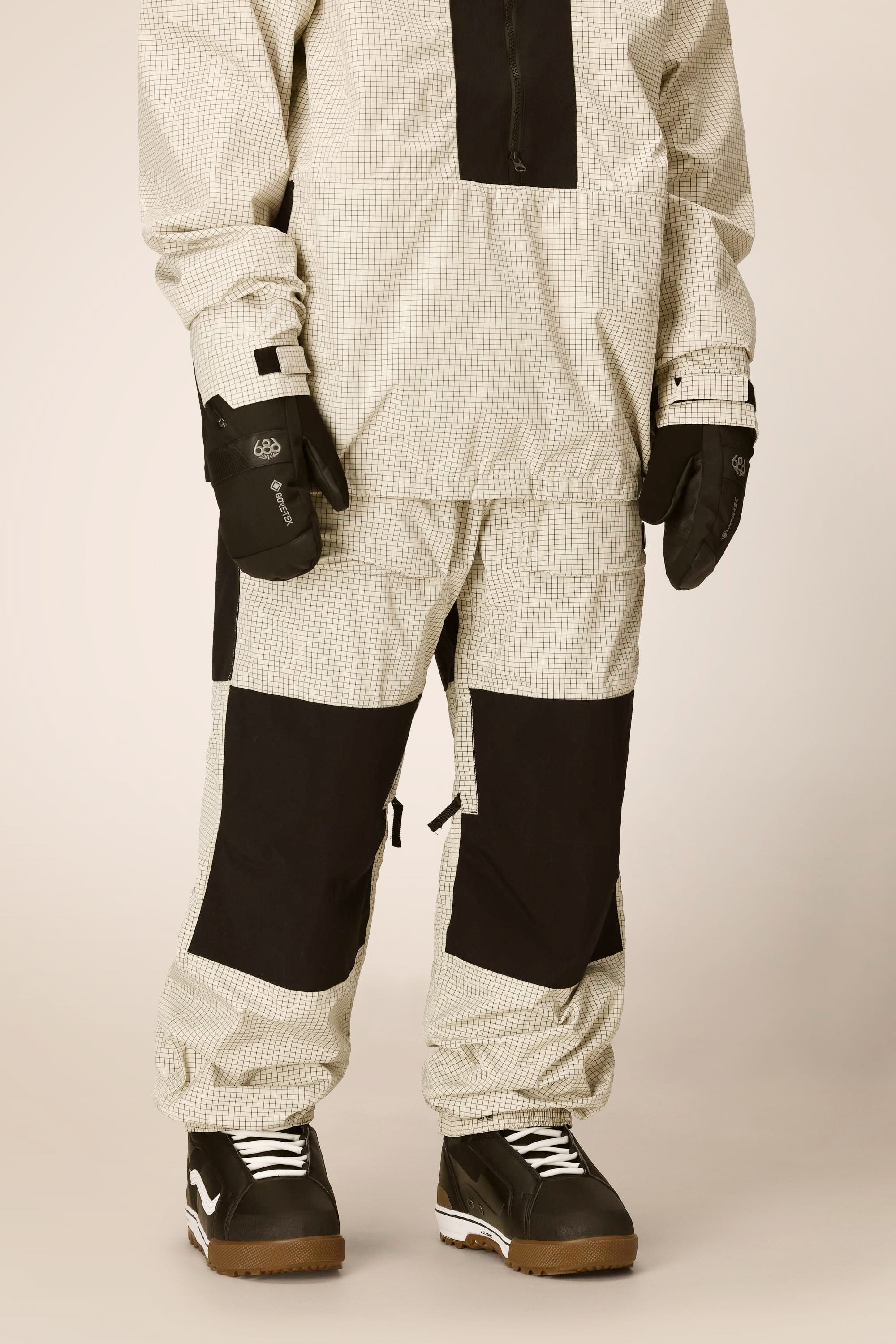 A person is wearing the 686 Ghost™ 2.5L Pant by 686 with a matching checkered beige and black snow jacket, black gloves, and black-and-white snow boots, standing against a plain background.