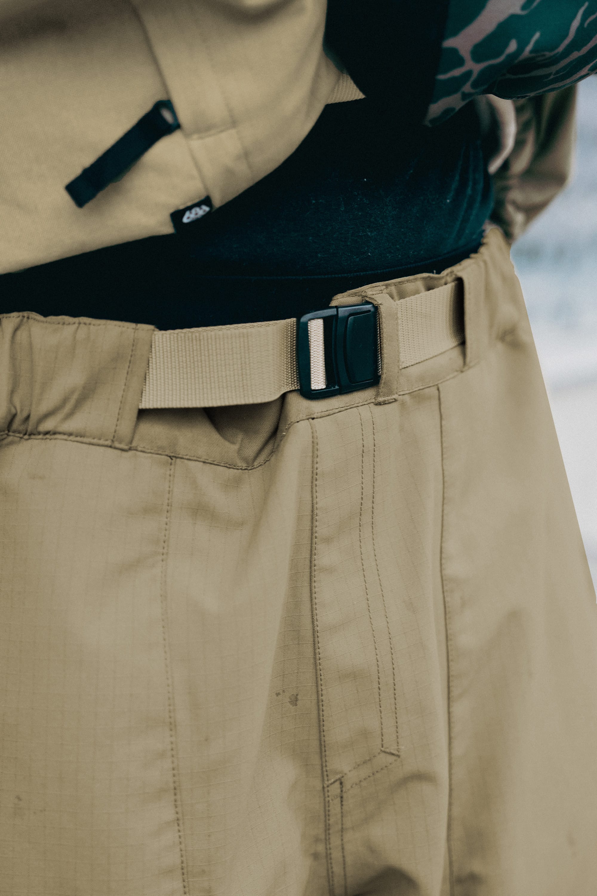 Close-up of a person wearing 686 Ghost™ 2.5L Pant by 686 with an integrated adjustable belt, paired with a black shirt, showcasing the pant’s beige flexible fabric, texture, and detailed stitching.