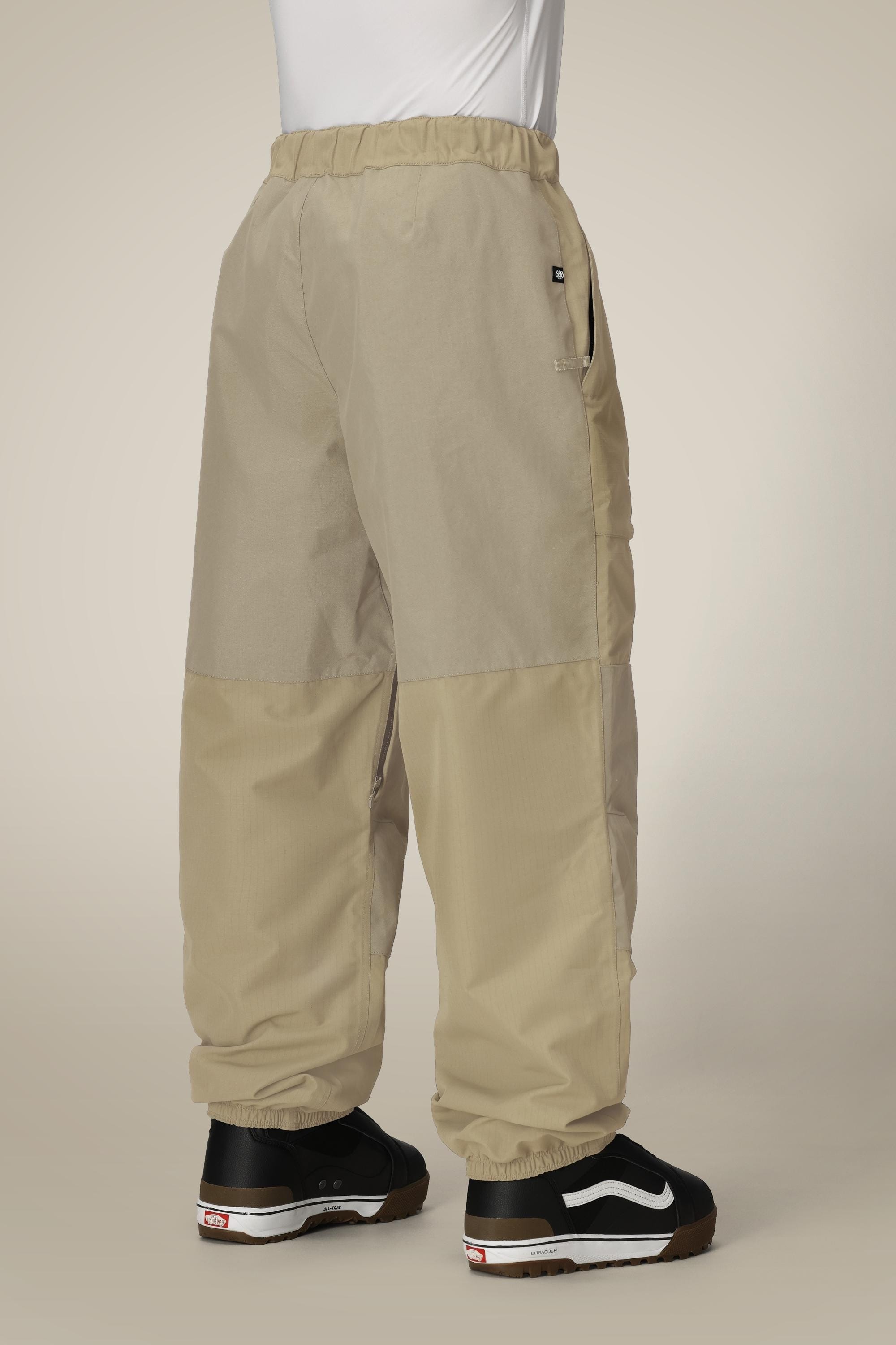 A person stands against a plain light background, seen from behind in relaxed-fit 686 Ghost™ 2.5L Pant by 686—loose-fitting, beige two-tone shell pants with elastic cuffs and waistband—paired with black and white sneakers.