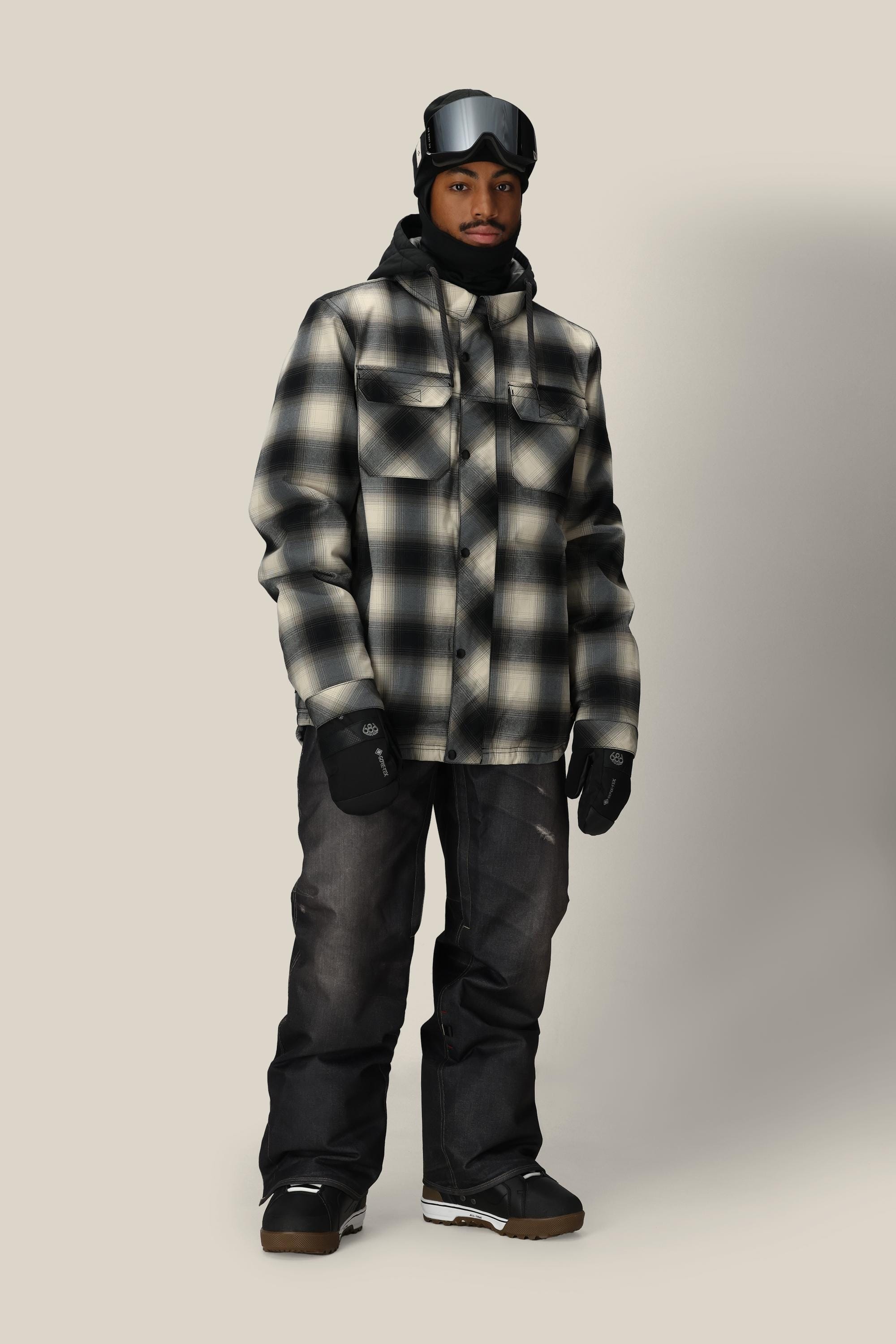A person stands against a plain background wearing winter attire, including the 686 Men's Deconstructed™ Denim Pant by 686, a black and white plaid jacket, gloves, goggles, and a beanie.
