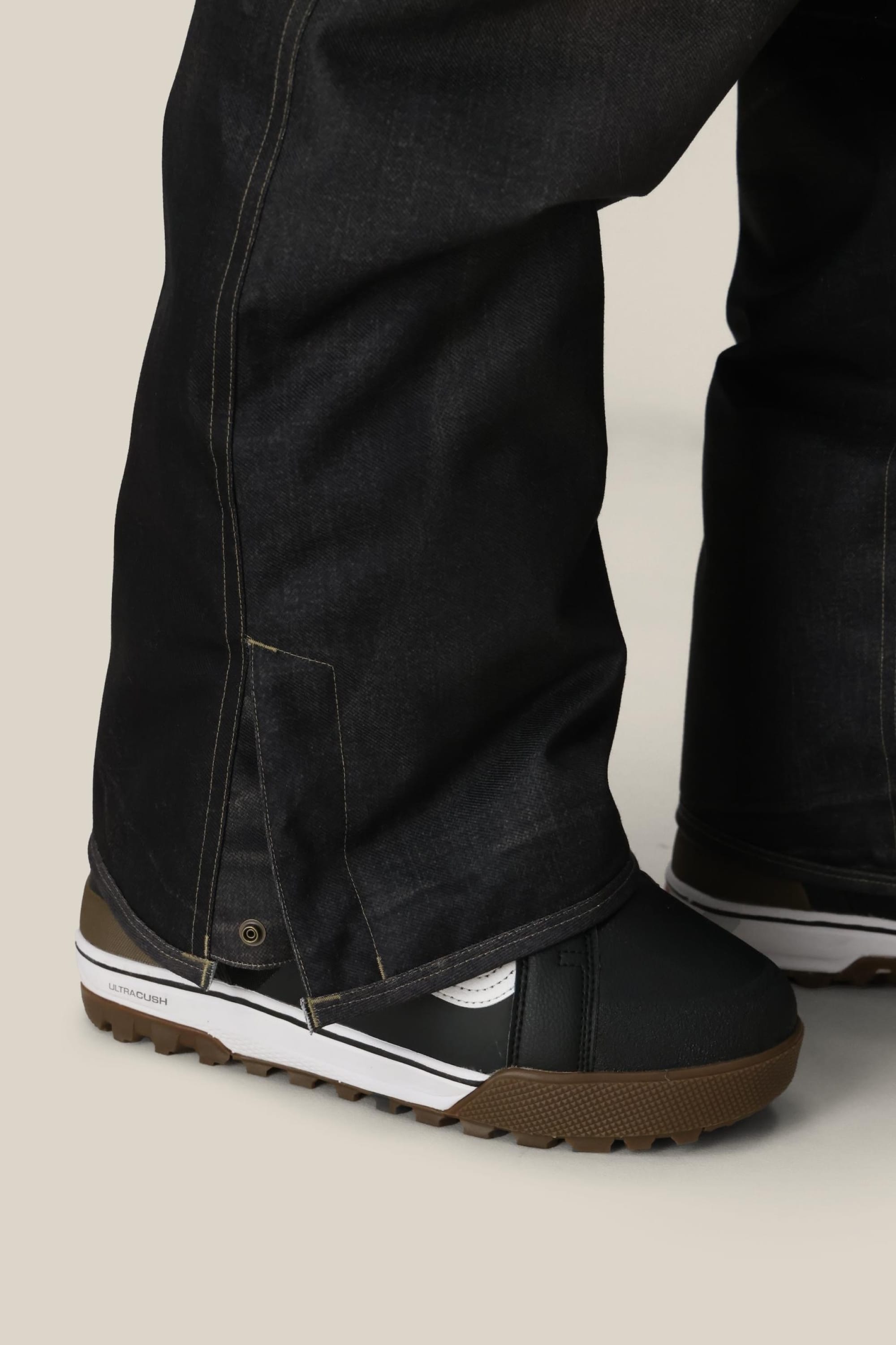 Close-up of a person wearing 686 Men's Deconstructed™ Denim Pant by 686, the wide-leg style covering black and white shoes with brown soles, standing on a light-colored floor.