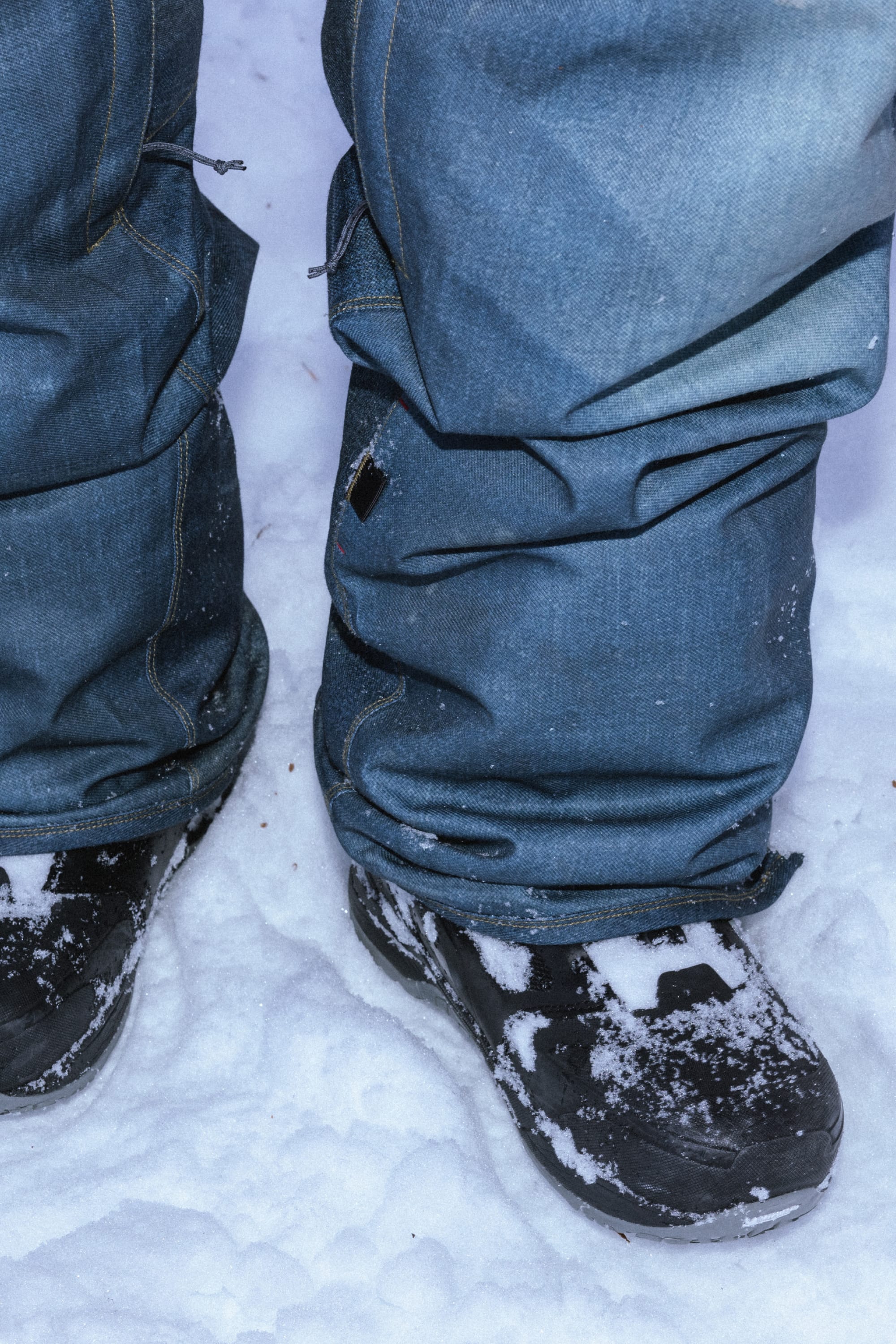 Close-up of a person shredding in the 686 Men's Deconstructed™ Denim Pant, with black snow boots and snow clinging to the boots and pant hems.