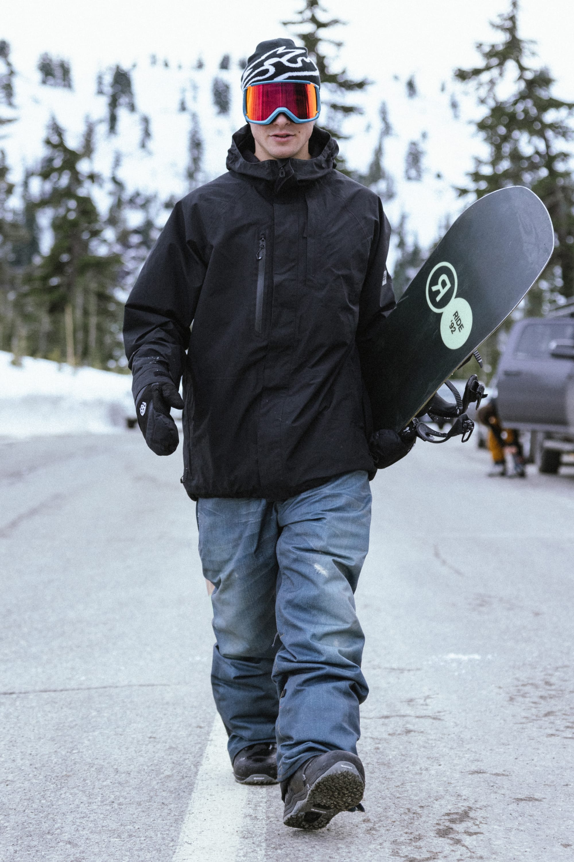 A snowboarder in a black jacket, 686 Men's Deconstructed™ Denim Pant by 686, and goggles walks on a snowy road carrying a snowboard, with snow-covered trees and parked cars behind him.