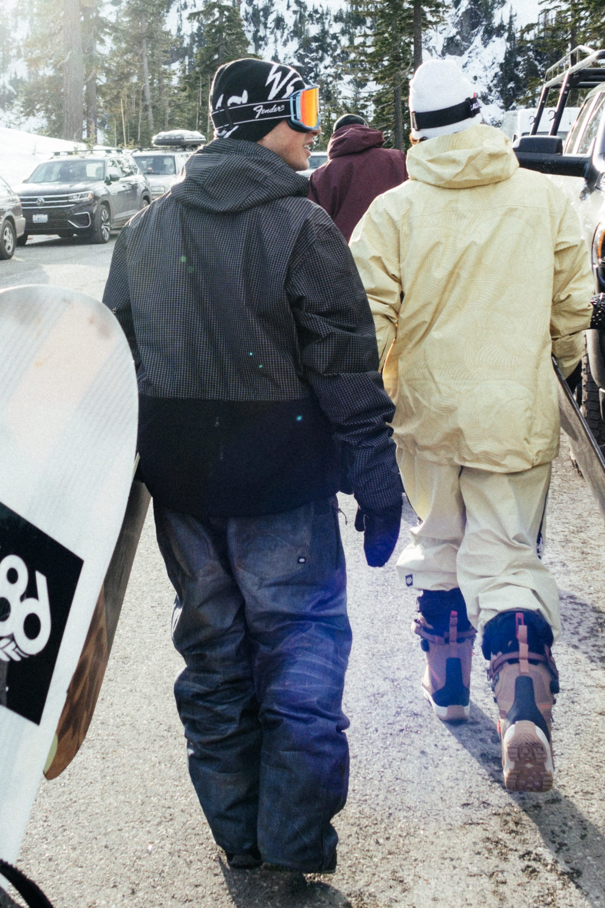 Two people in winter gear walk a snow-lined road, carrying snowboards and rocking 686 Men's Deconstructed™ Denim Pants. Parked cars, tall pines, and snowy mountains set the scene for an epic day on the slopes. Brand: 686.