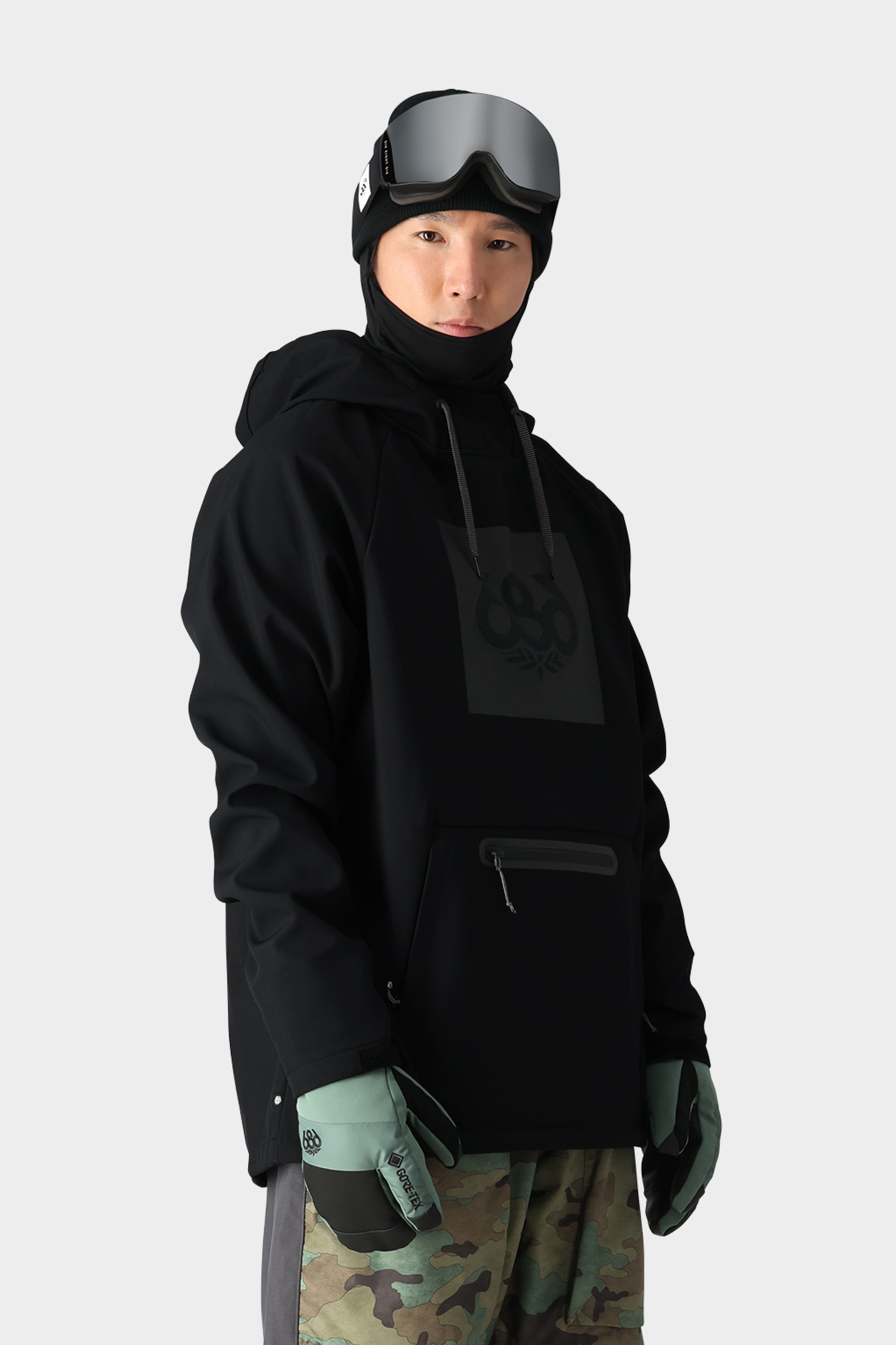 A person stands against a plain light gray background wearing the 686 Mens Waterproof Hoody in black, paired with a black balaclava, ski goggles, mint green gloves, and camouflage pants.