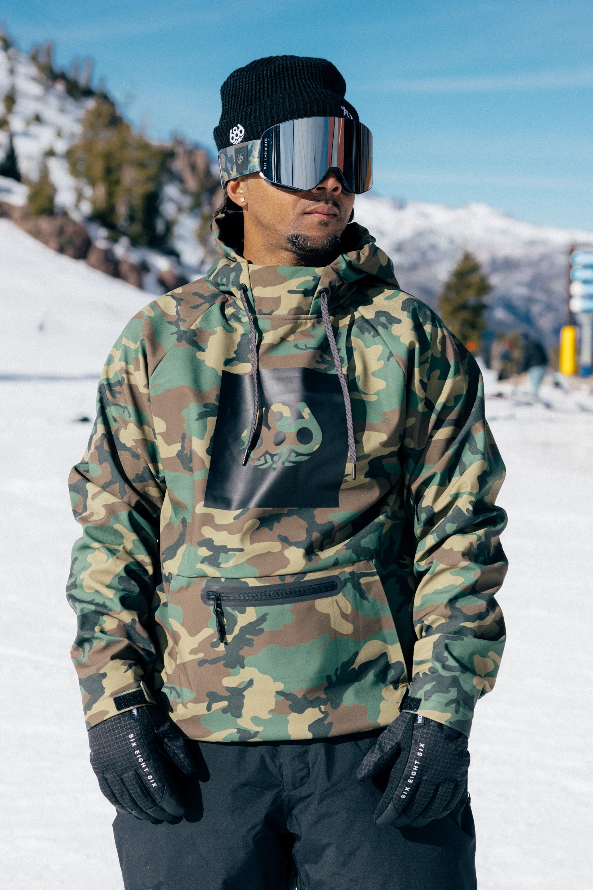 A man wearing the 686 Men’s Waterproof Hoody by 686, along with black gloves, a beanie, and ski goggles, stands on a snowy mountain with trees and distant peaks under a blue sky.