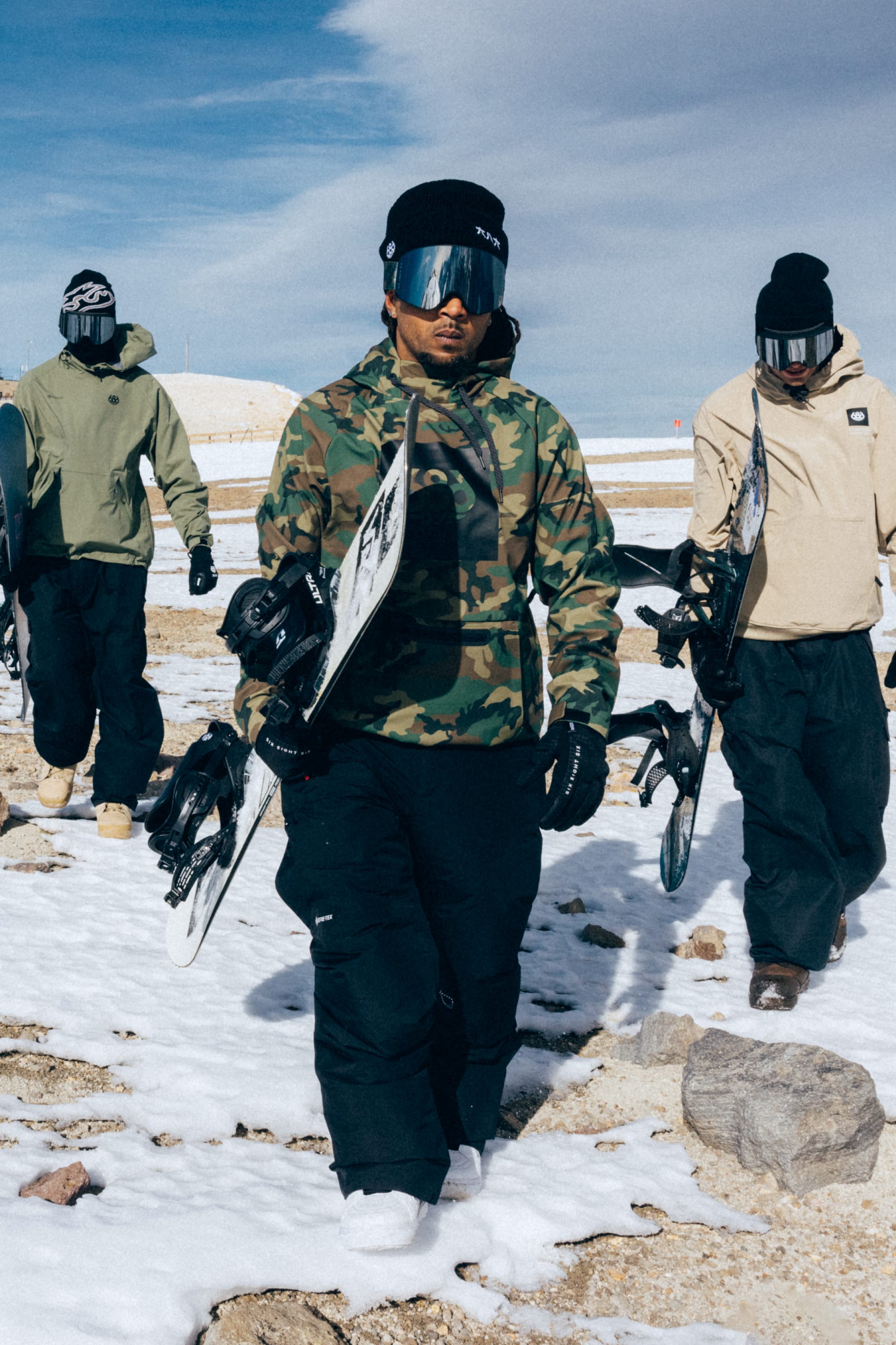 Three snowboarders in winter gear walk across the snow, each carrying a snowboard. The person in front is wearing a 686 Men’s Waterproof Hoody by 686 with black pants. Behind them stretches a snowy, rocky landscape beneath a partly cloudy sky.