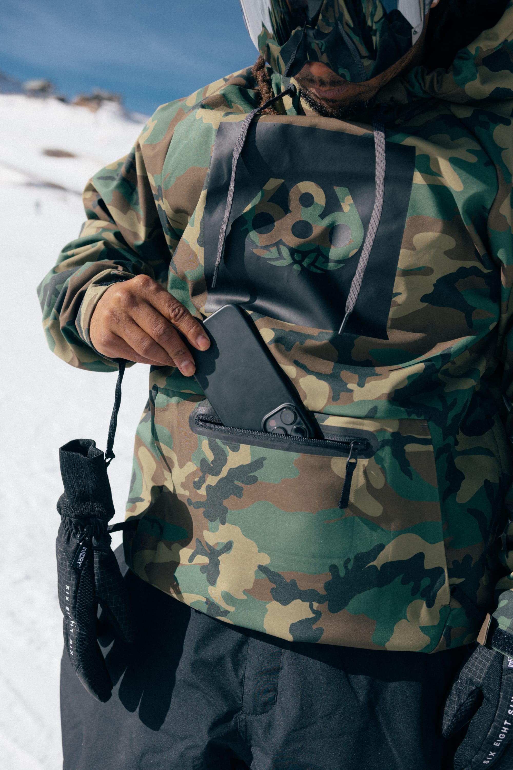 Wearing the 686 Men’s Waterproof Hoody by 686, a person dressed in ski gear with black gloves slips a smartphone into the front pocket while standing on snowy ground; their face remains unseen.