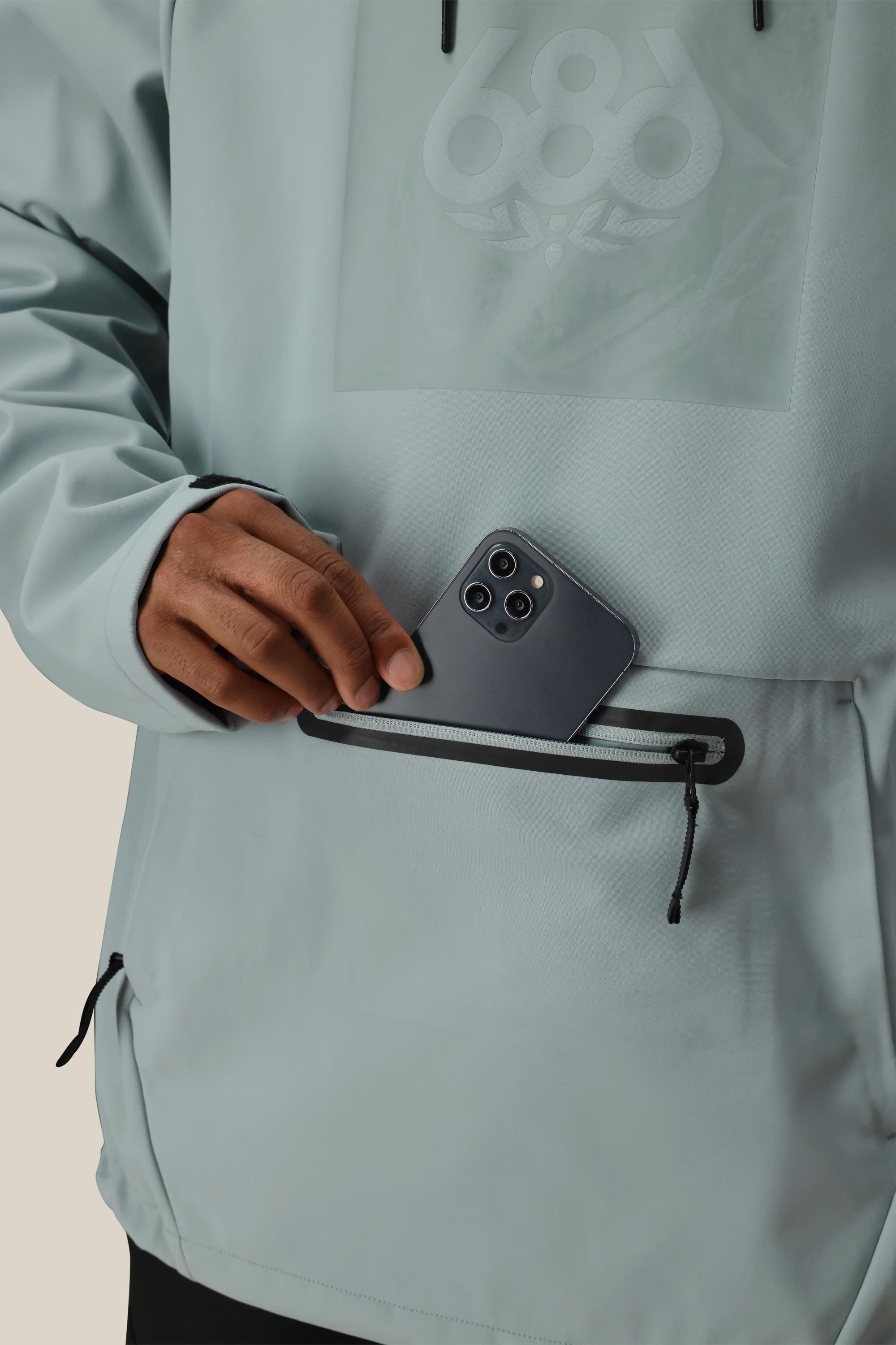 A person wearing the 686 Mens Waterproof Hoody in light gray places a black smartphone into its zippered front pocket.