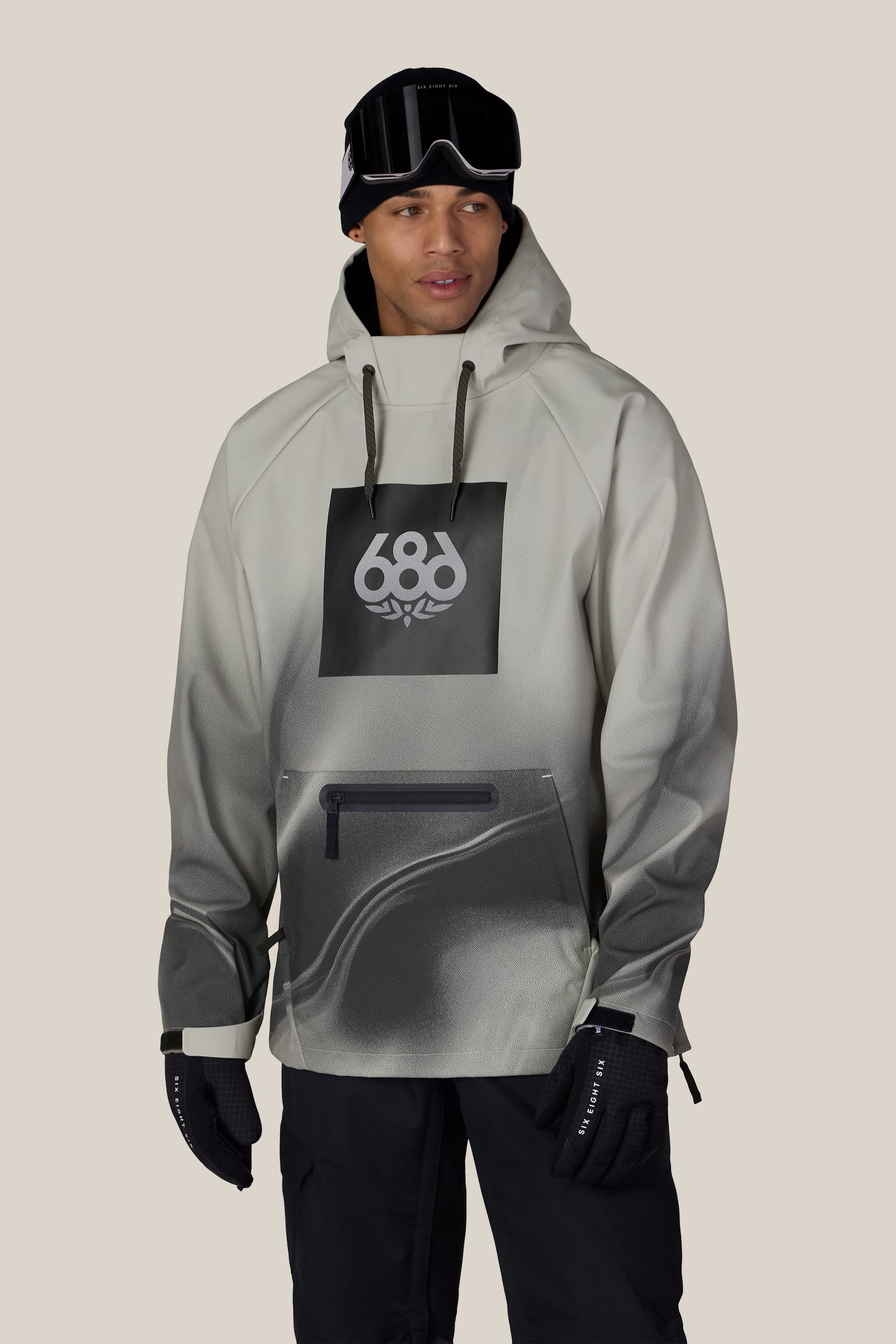 A person wearing the 686 Men’s Waterproof Hoody in a gray gradient color, featuring the 686 logo, along with black gloves, black snow pants, a black beanie, and ski goggles, standing against a plain light background.