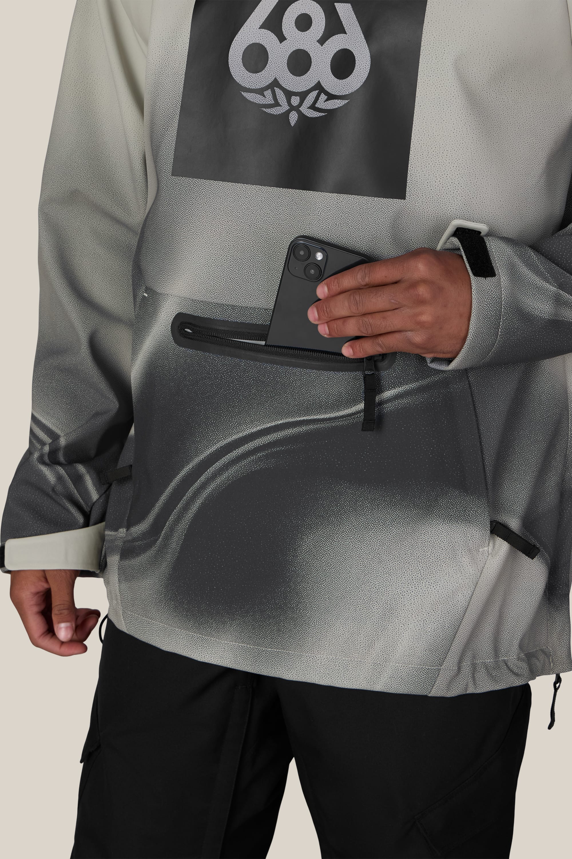 A person wearing the 686 Men’s Waterproof Hoody, a gray-and-black jacket by 686, is placing a black smartphone into the zippered front pocket. Only their lower torso and one hand are visible.