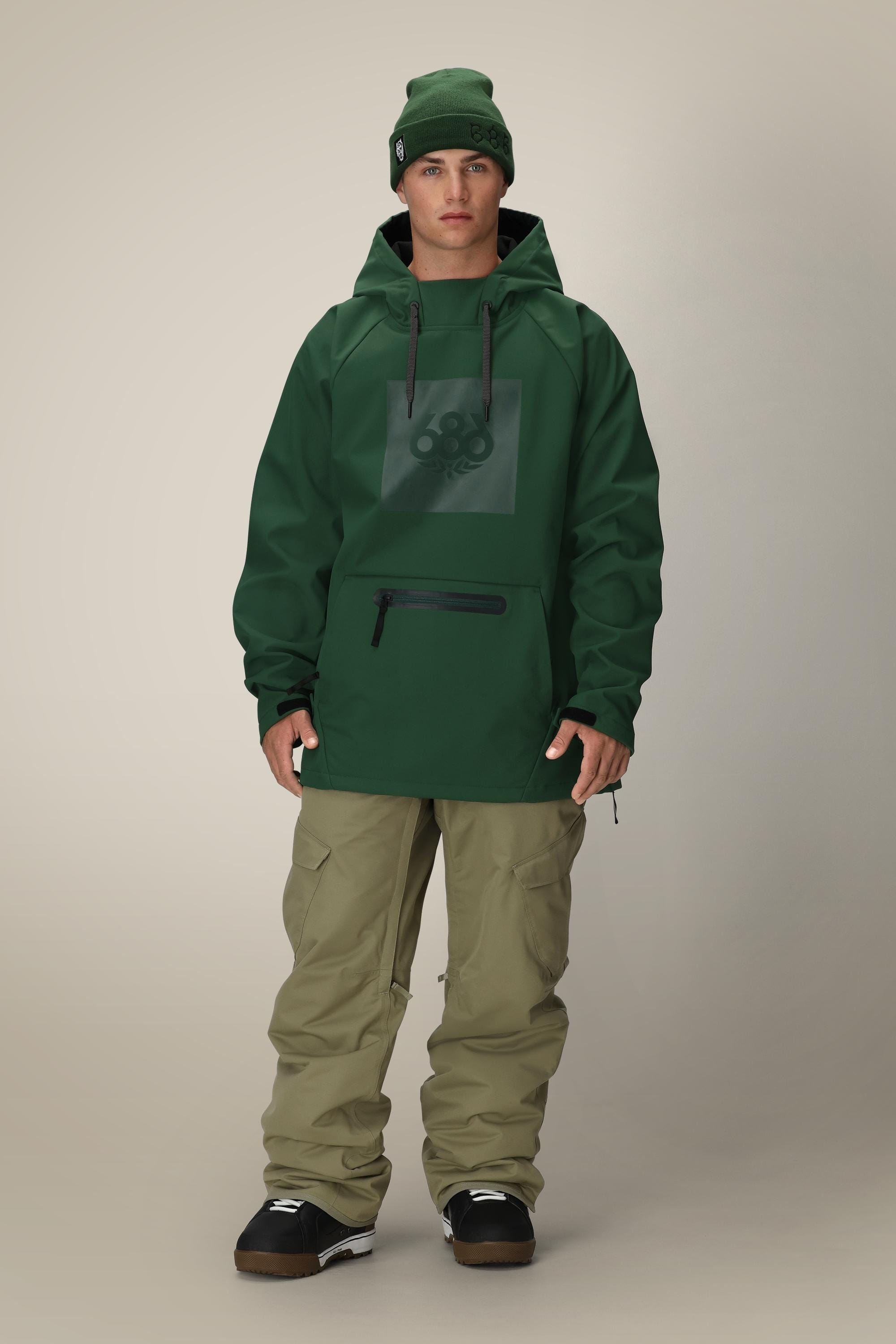 A person wears the 686 Mens Waterproof Hoody in dark green with a front pocket and subtle logo, paired with a matching beanie, khaki pants, and black snow boots, standing against a plain beige background.