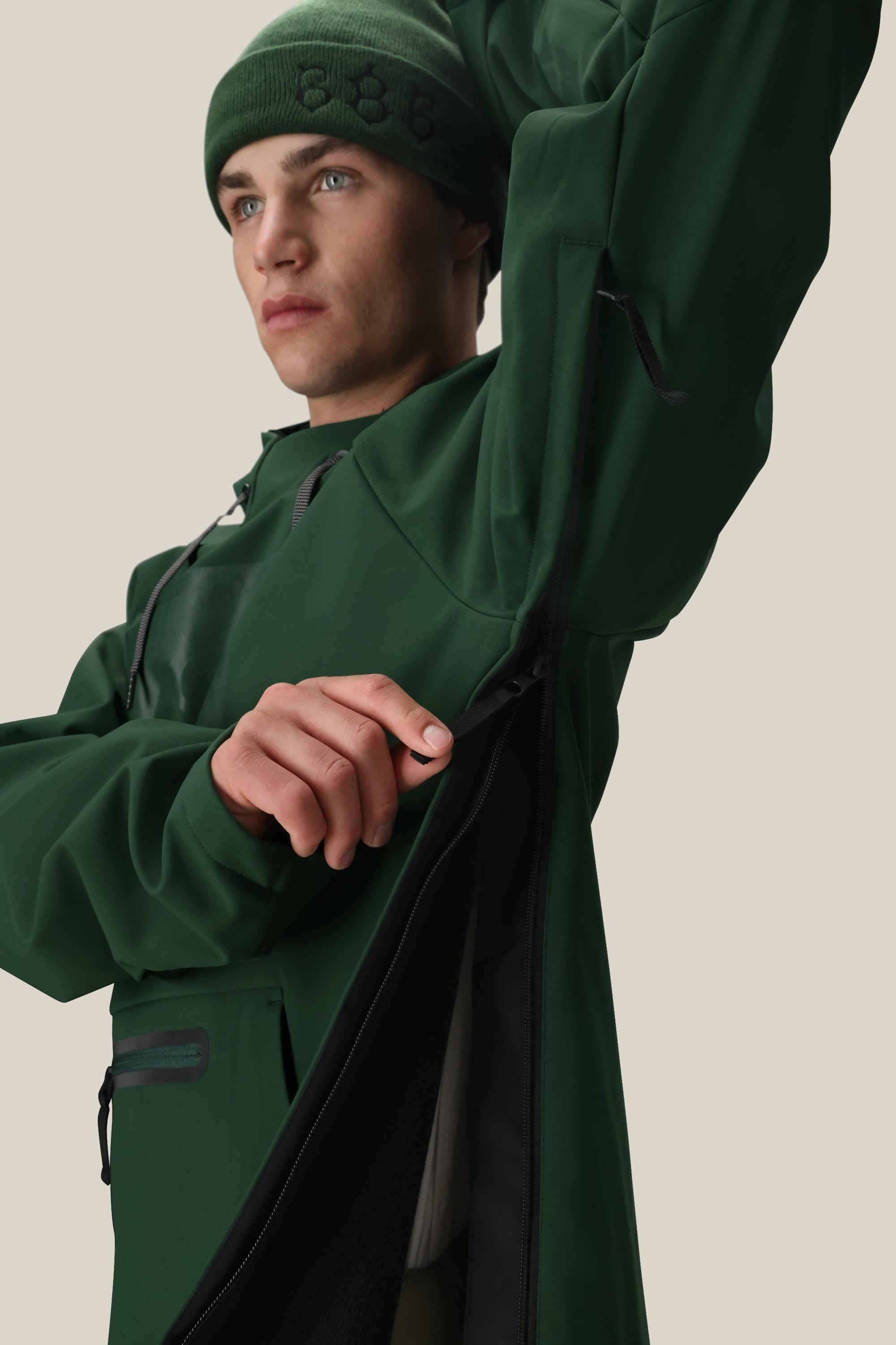A person wearing the 686 Men’s Waterproof Hoody in green and a beanie opens the large side zipper under their arm, revealing a black inner lining. The background is light beige.