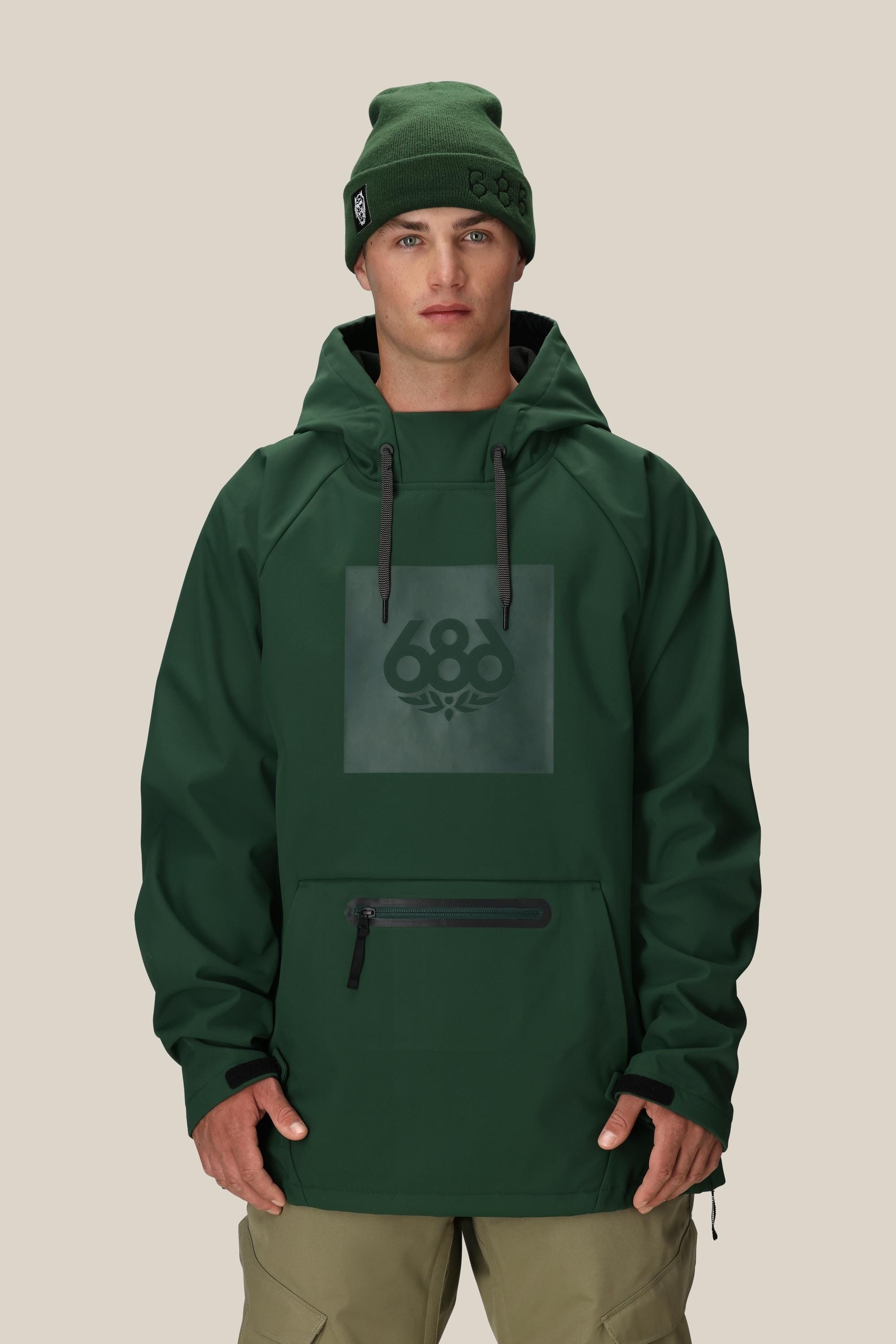A man faces forward against a beige background wearing the 686 Mens Waterproof Hoody in dark green, featuring a black zipper, front pocket, and fleece-lined interior, along with matching khaki pants and a green 686 beanie.