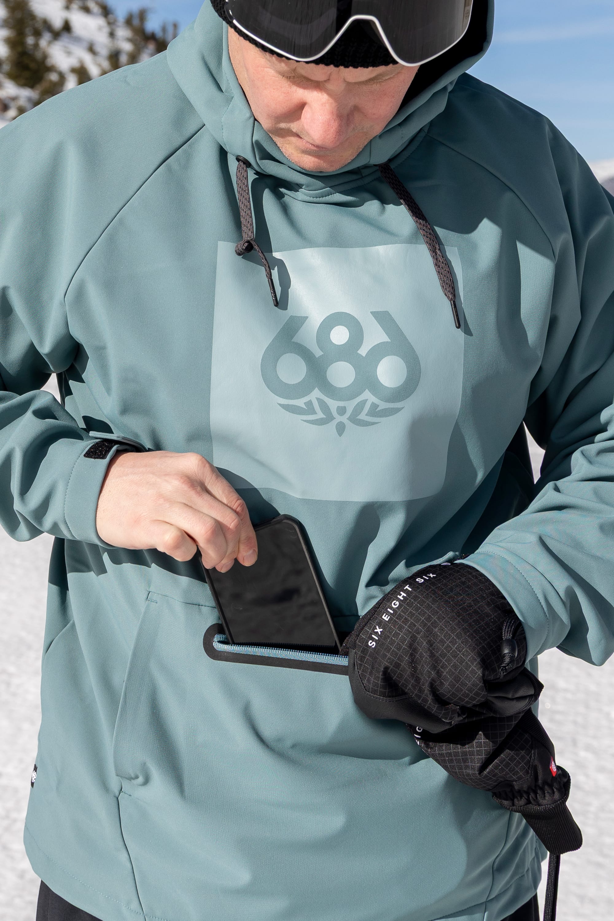 Wearing the 686 Men’s Waterproof Hoody, a person with black gloves stands on snow and puts a smartphone into the front zippered pocket.