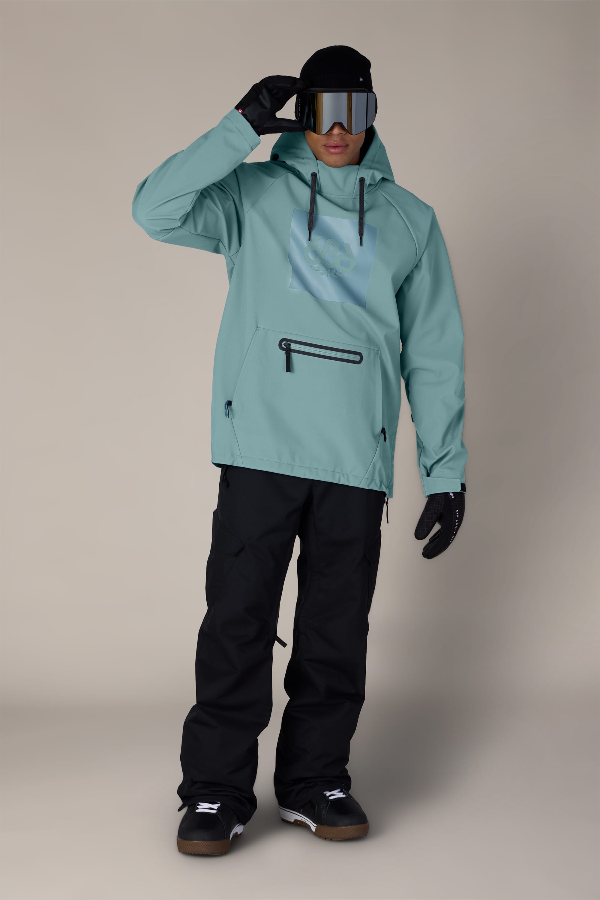 Wearing the 686 Men’s Waterproof Hoody in light blue, along with black snow pants, gloves, a beanie, and ski goggles, a person stands on a neutral background and raises one hand to adjust their goggles.