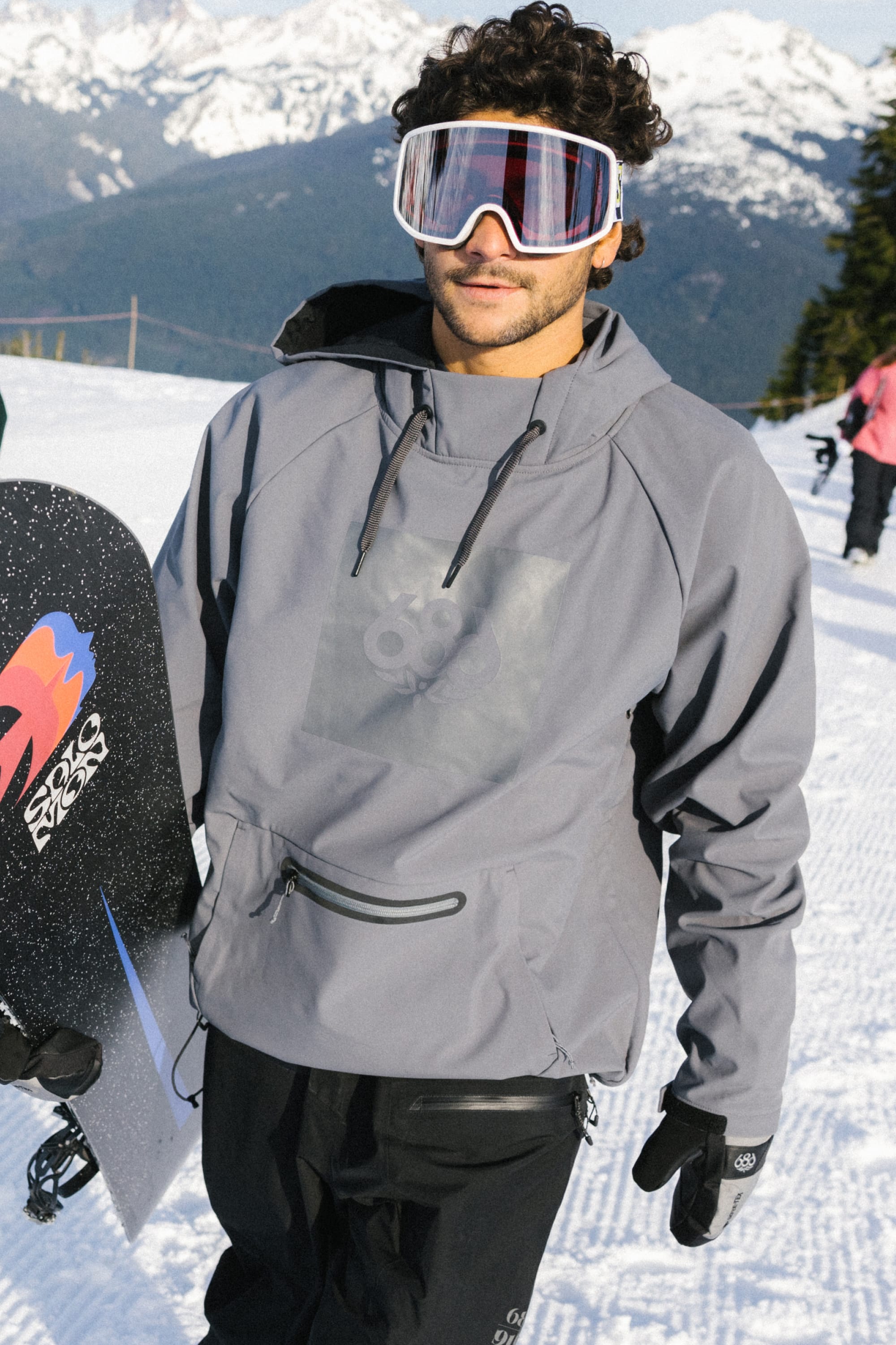 A man wearing the 686 Men’s Waterproof Hoody stands on a snowy mountain, holding a snowboard, with snow-covered trees and mountains visible in the background.