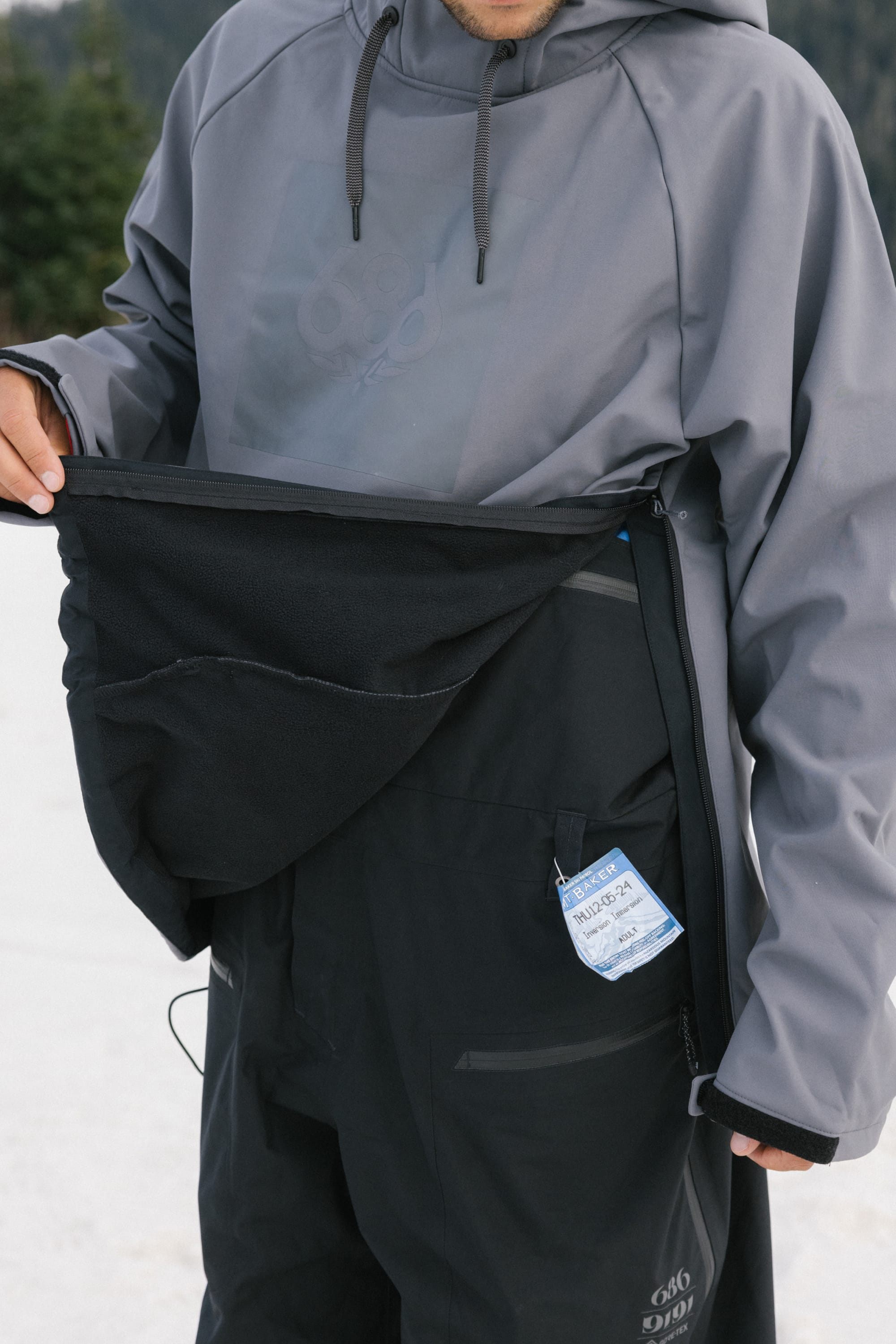 Wearing the 686 Mens Waterproof Hoody and black snow pants, a person stands outside amid snow and trees, displaying a large fleece-lined front pocket with a blue ski pass tucked inside.
