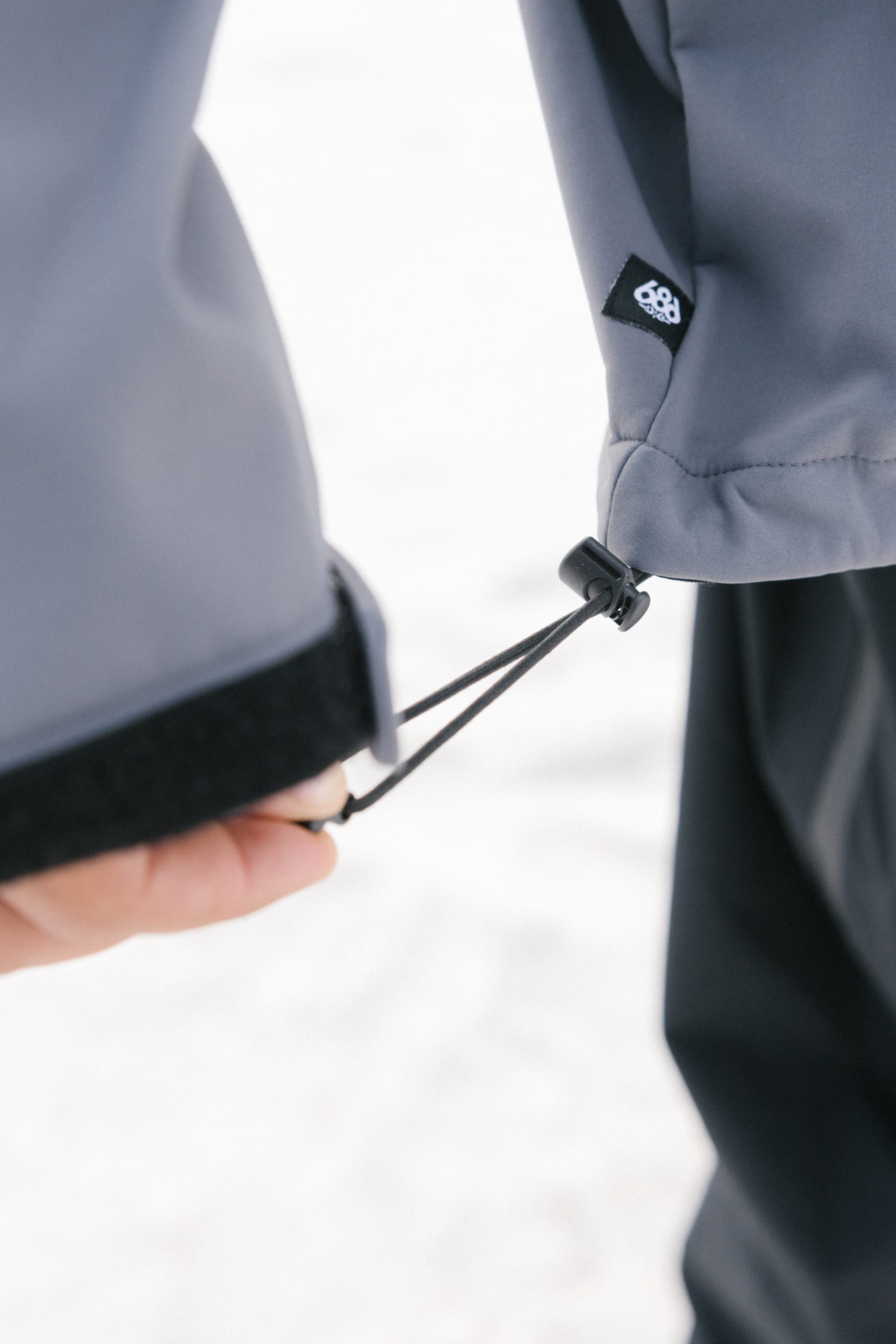 A close-up shows someone adjusting the elastic cuff drawstring on a gray 686 Mens Waterproof Hoody sleeve, fleece lined with a visible black and white 686 logo. The background is blurred and light-colored.