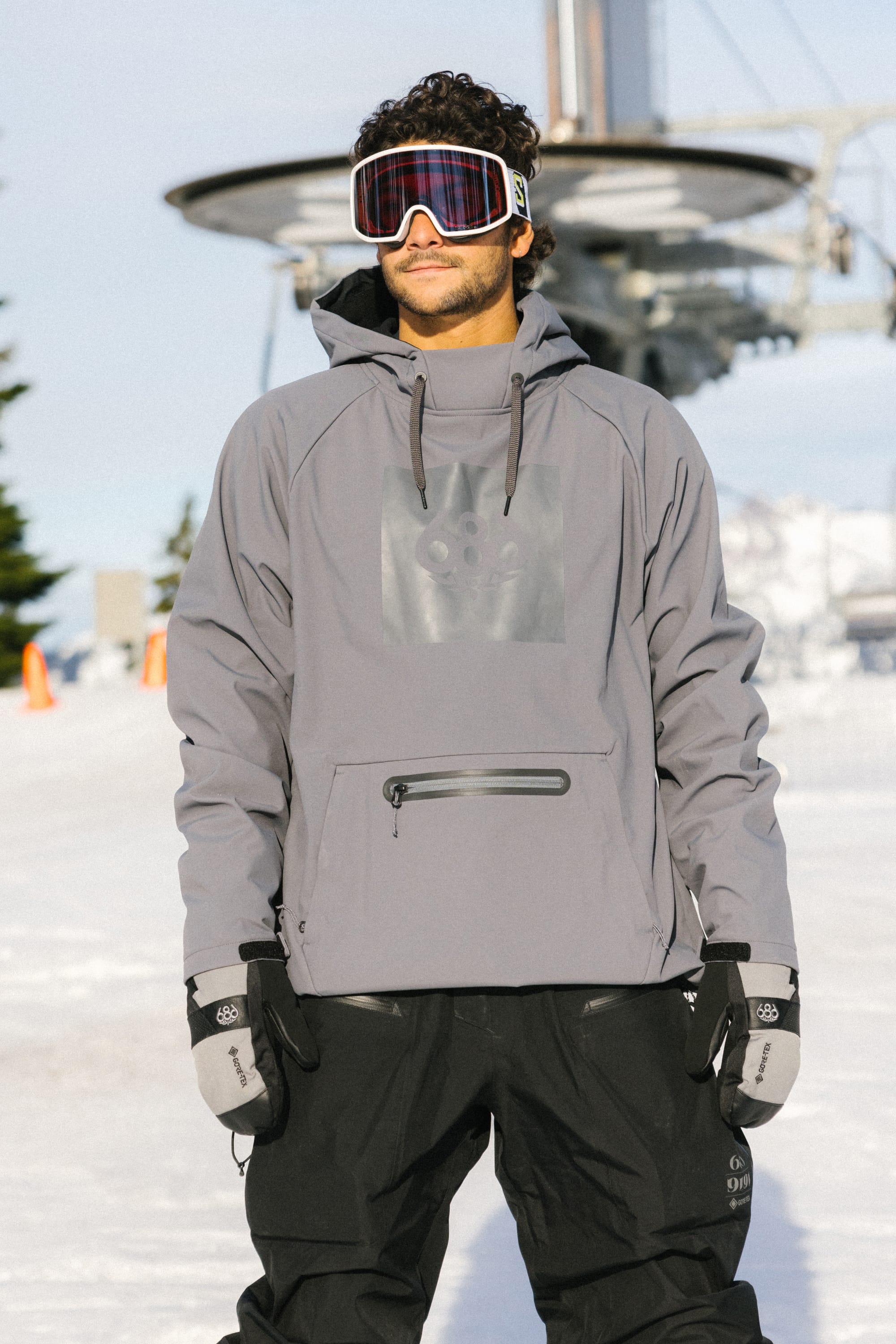A person wearing the 686 Men’s Waterproof Hoody by 686, along with black ski pants, gloves, and ski goggles, stands on snow with a ski lift and mountains in the background.