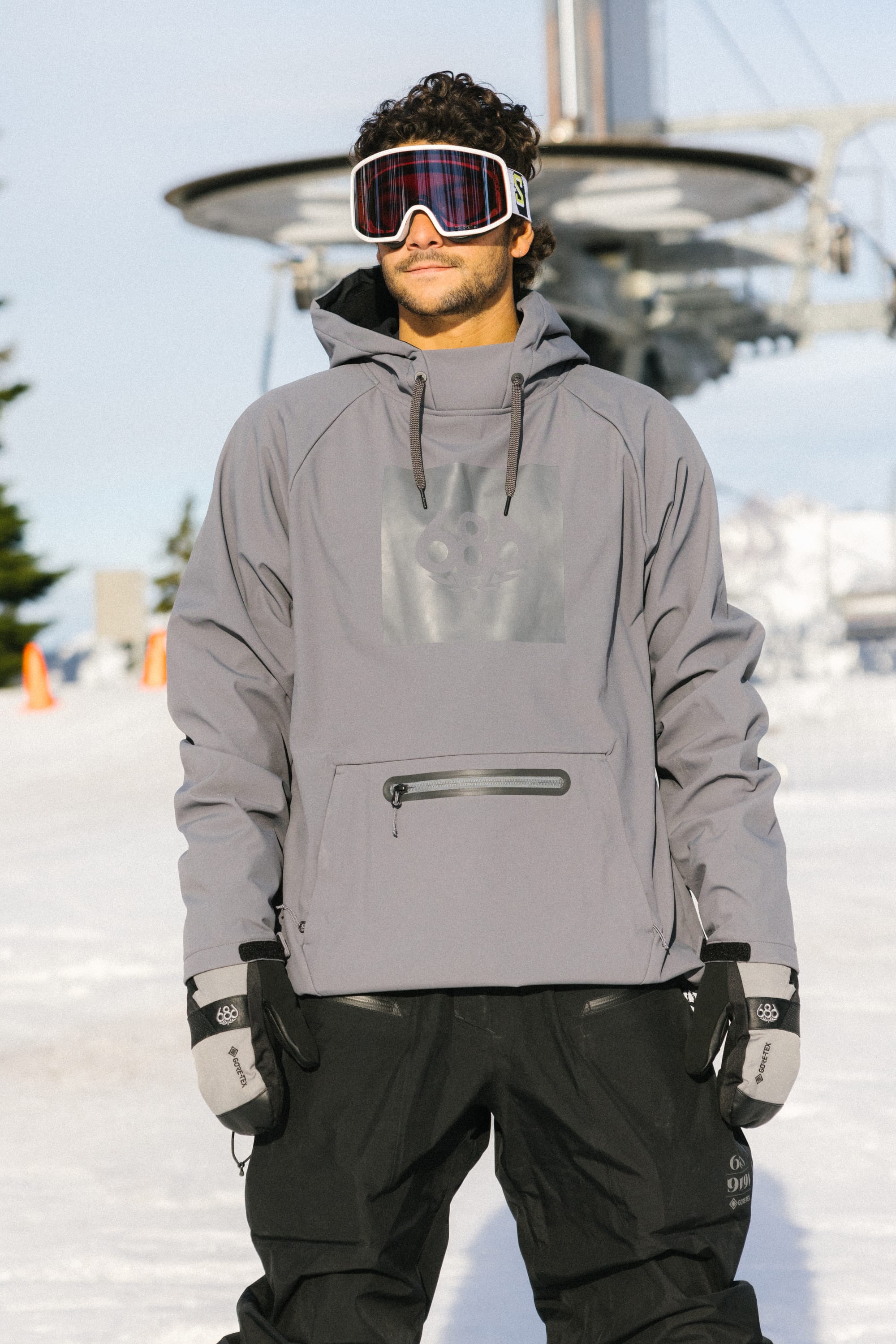 A man wearing a 686 Mens Waterproof Hoody stands on a snowy slope with black snow pants, gloves, ski goggles, a ski lift, and mountains in the background.